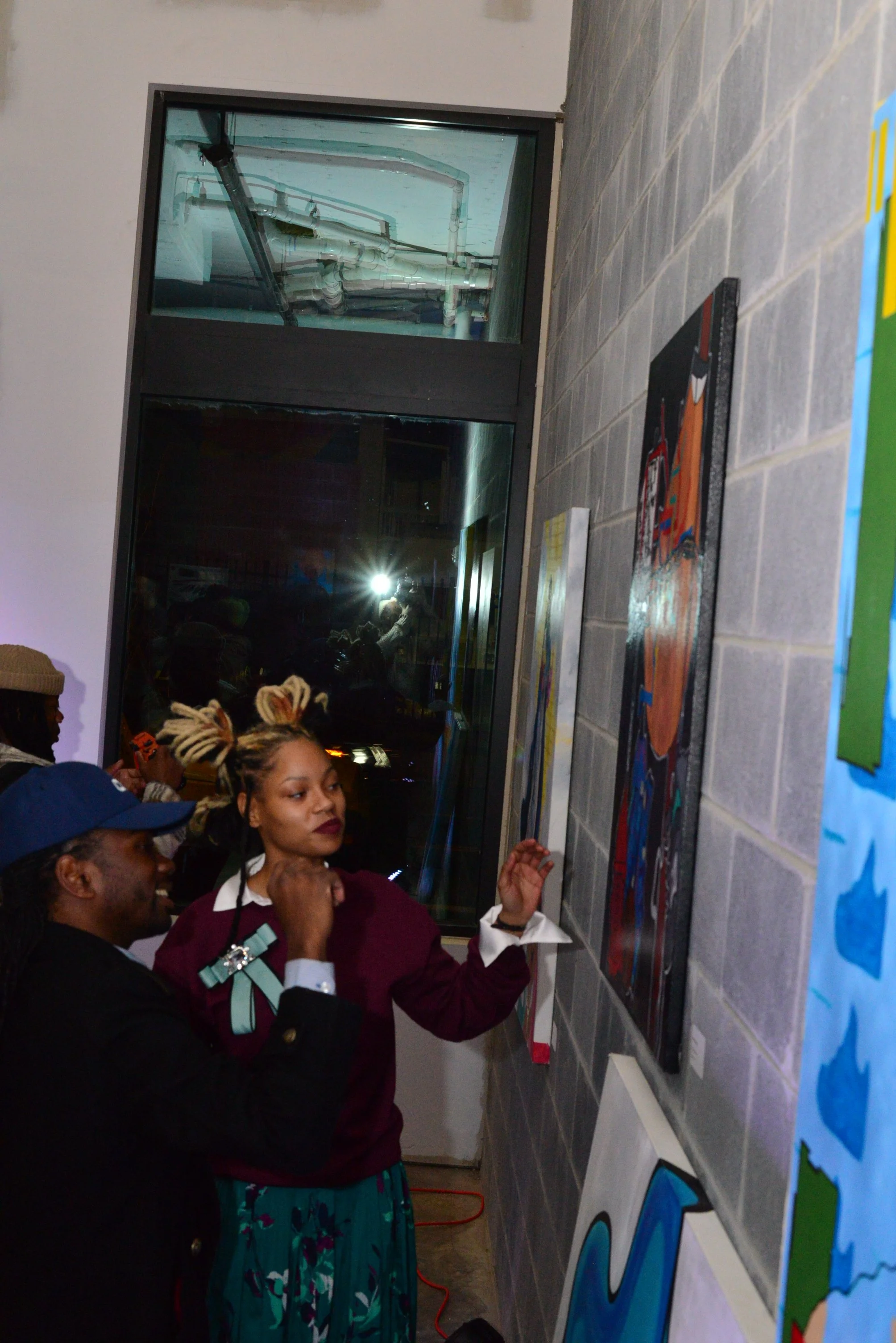 A group of people gathered in an art gallery, looking at colorful paintings on a gray brick wall. The room has a large window showing pipes and utility structures outside, and reflections of people are visible in the glass.