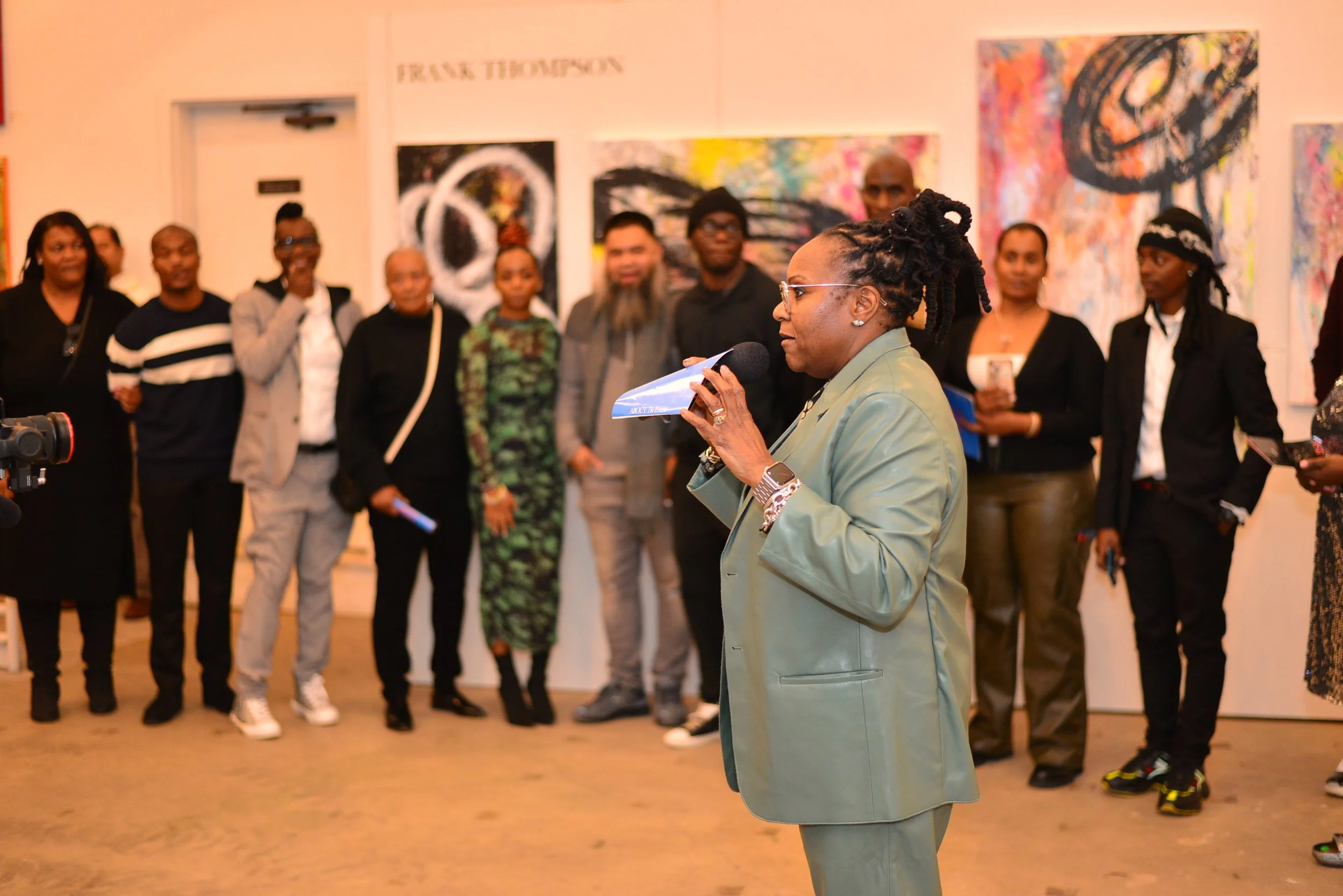 An older woman with glasses and dreadlocks speaking into a microphone at an art gallery event, with a group of diverse people and colorful abstract paintings in the background.