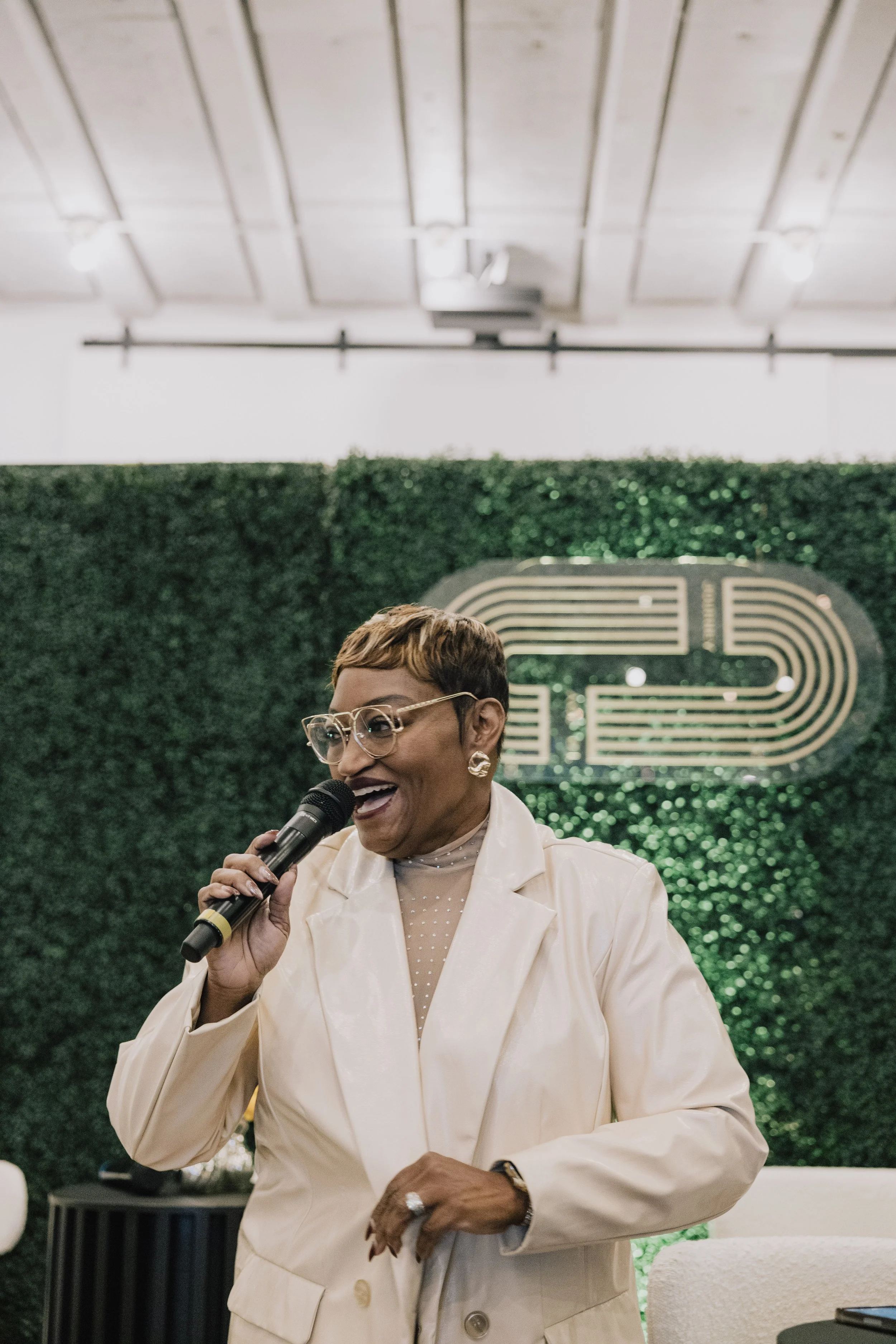 A woman with short hairstyle, glasses, and earrings, dressed in a white suit, smiling and speaking into a microphone at an indoor event with green foliage and a decorative emblem in the background.