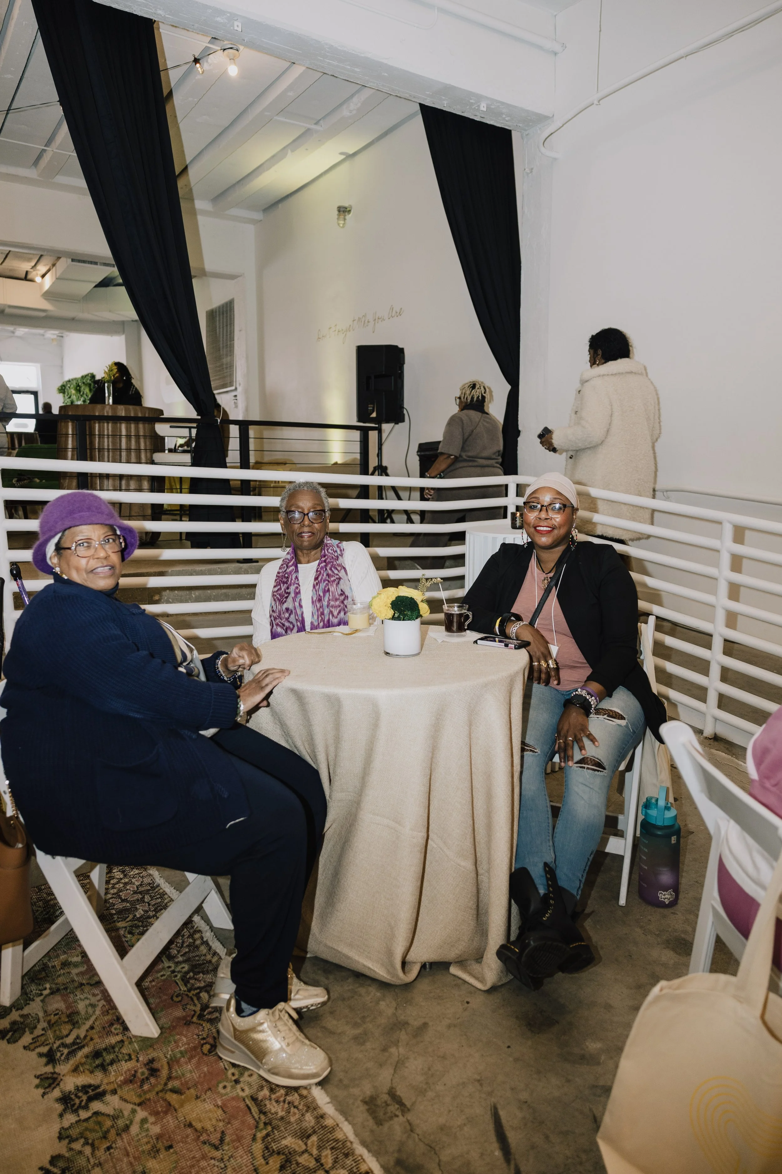 Three women sitting at a round banquet table with a beige tablecloth in a spacious, industrial-style event space. Two women in foreground wearing glasses, one with a purple hat, the other with a headscarf, smiling at the camera. The third woman on the right wears glasses and a pink top. There are decorations including a small potted plant with yellow flowers on the table. In the background, two women are standing, one with a fur coat and the other with short hair and glasses, with a stage area partially visible behind black curtains.
