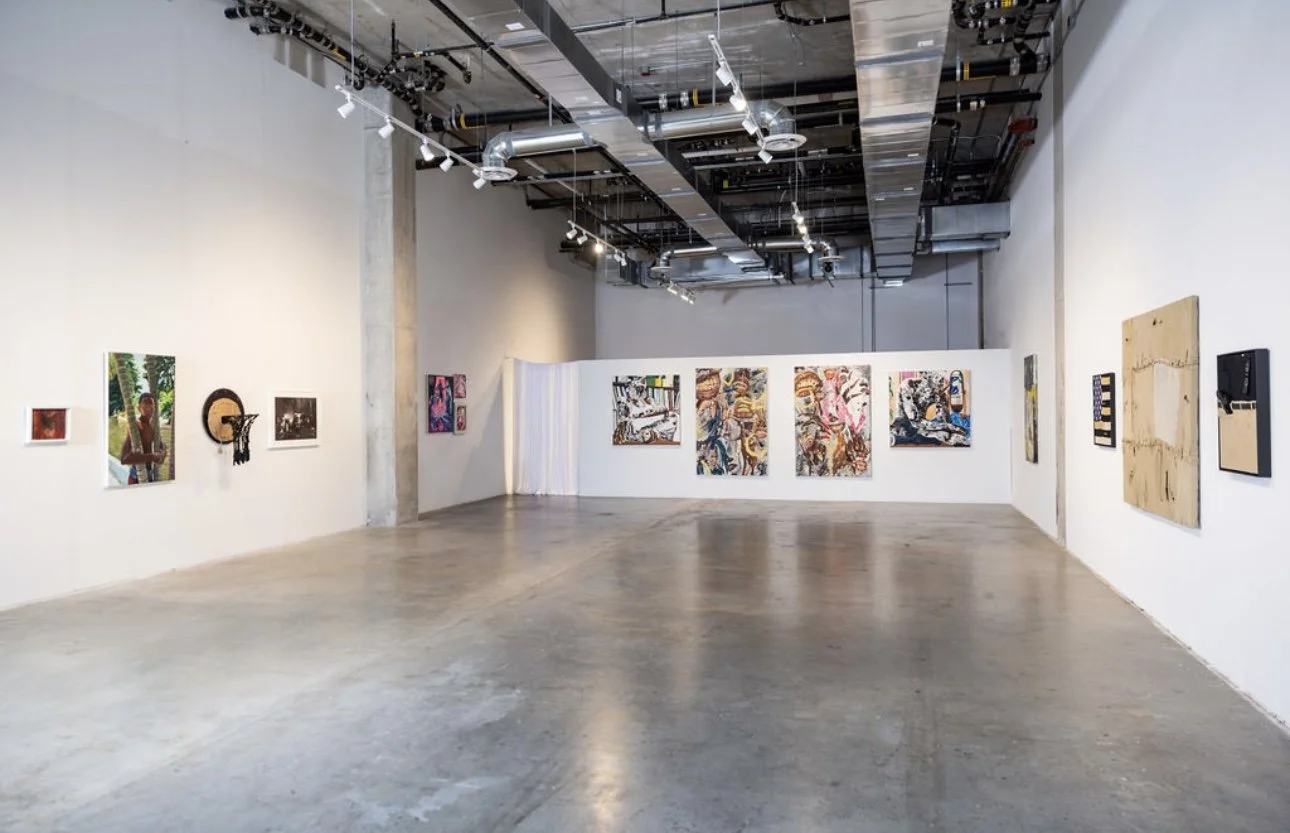 Interior view of an art gallery with white walls and artwork displayed in a row, including paintings, mixed media, and photographs, with industrial ceiling and polished concrete floor.