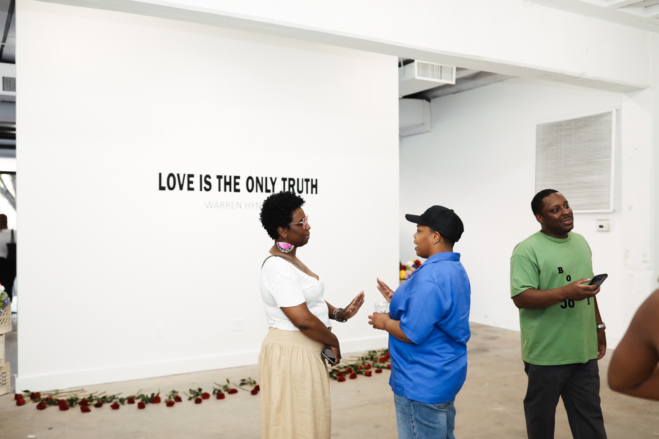 Three people are engaged in conversation in front of a white wall with a quote by Warren H. in black letters reading "LOVE IS THE ONLY TRUTH." There is a flower arrangement on the floor at the base of the wall. The individuals are wearing casual clothing, and one person on the right is holding a phone.