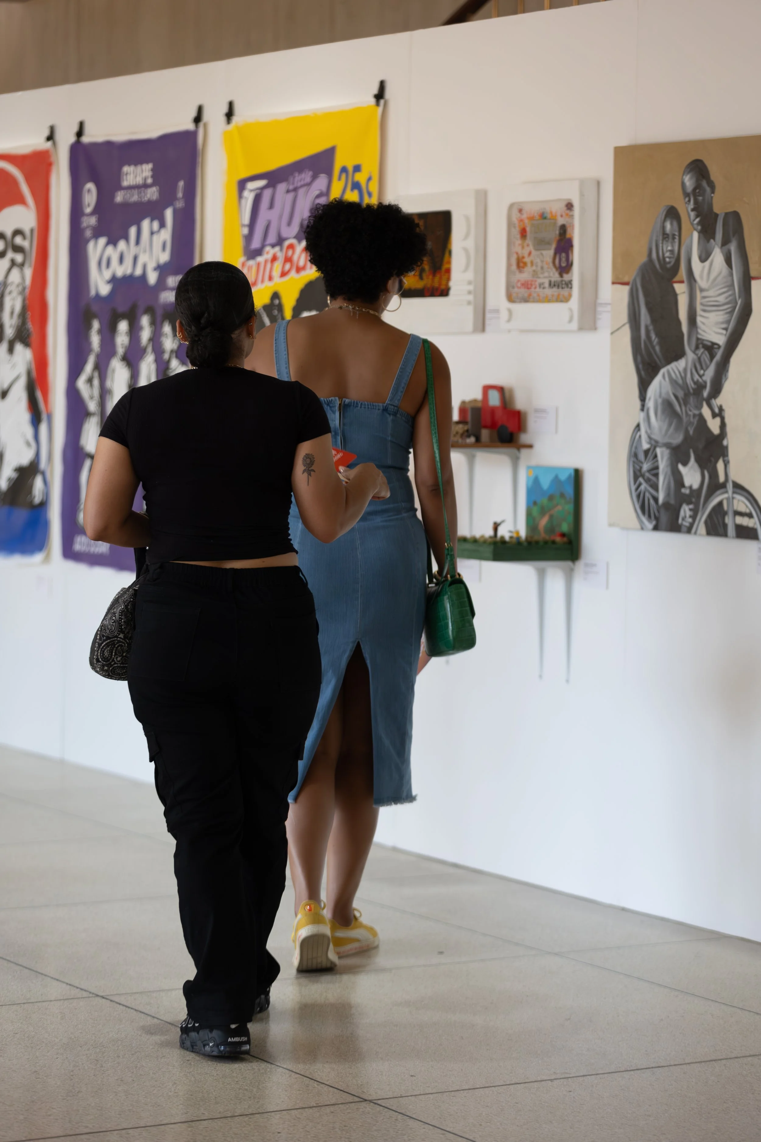 Two women walking in an art gallery. One in black, the other in a denim dress. Behind them are colorful posters on the wall and a small display shelf.