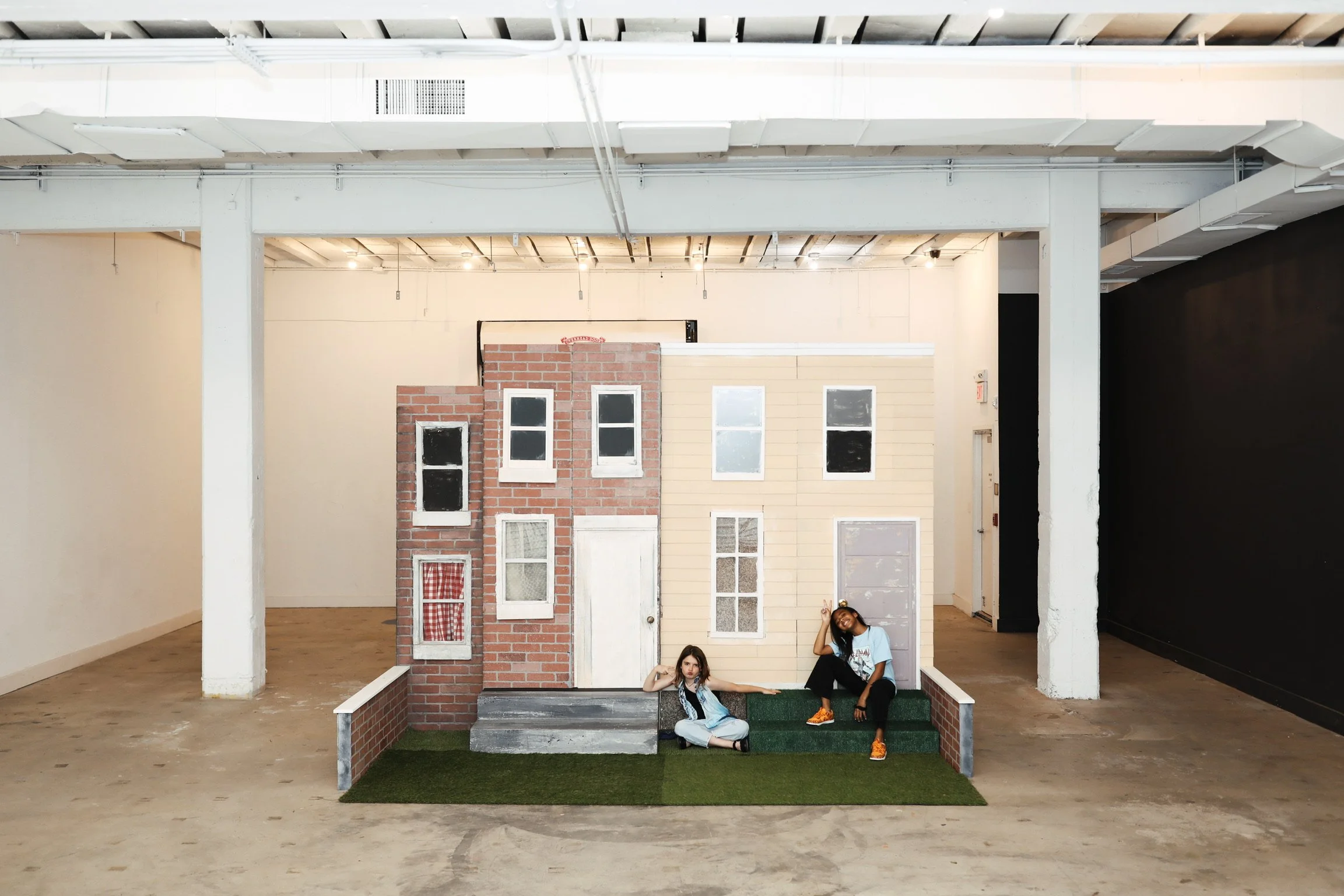 Two young women sitting on steps in front of a small set with cardboard and brick facade, resembling a miniature house, in an indoor space with exposed ceiling and large white columns.