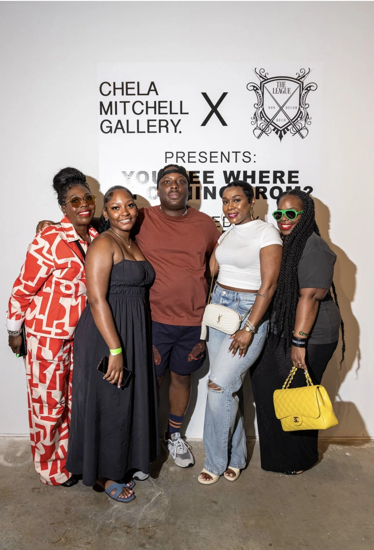 Group of five people standing together in front of a white wall with a poster. The poster reads Chela Mitchell Gallery presents: Who Are You Coming From?