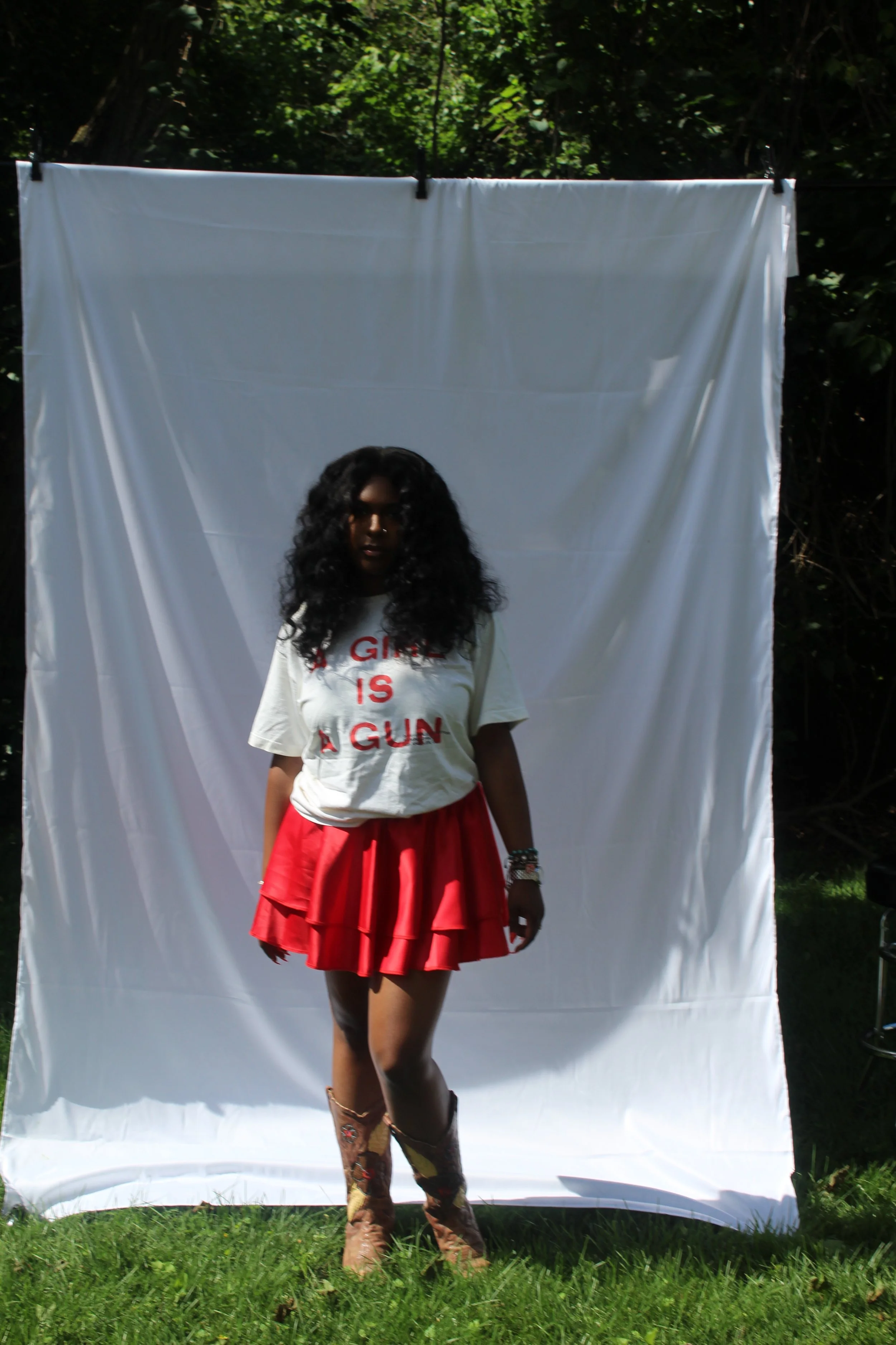 A woman standing outdoors in front of a white backdrop, wearing a white T-shirt with red text, a red skirt, cowboy boots, and accessories, with trees and grass surrounding her.
