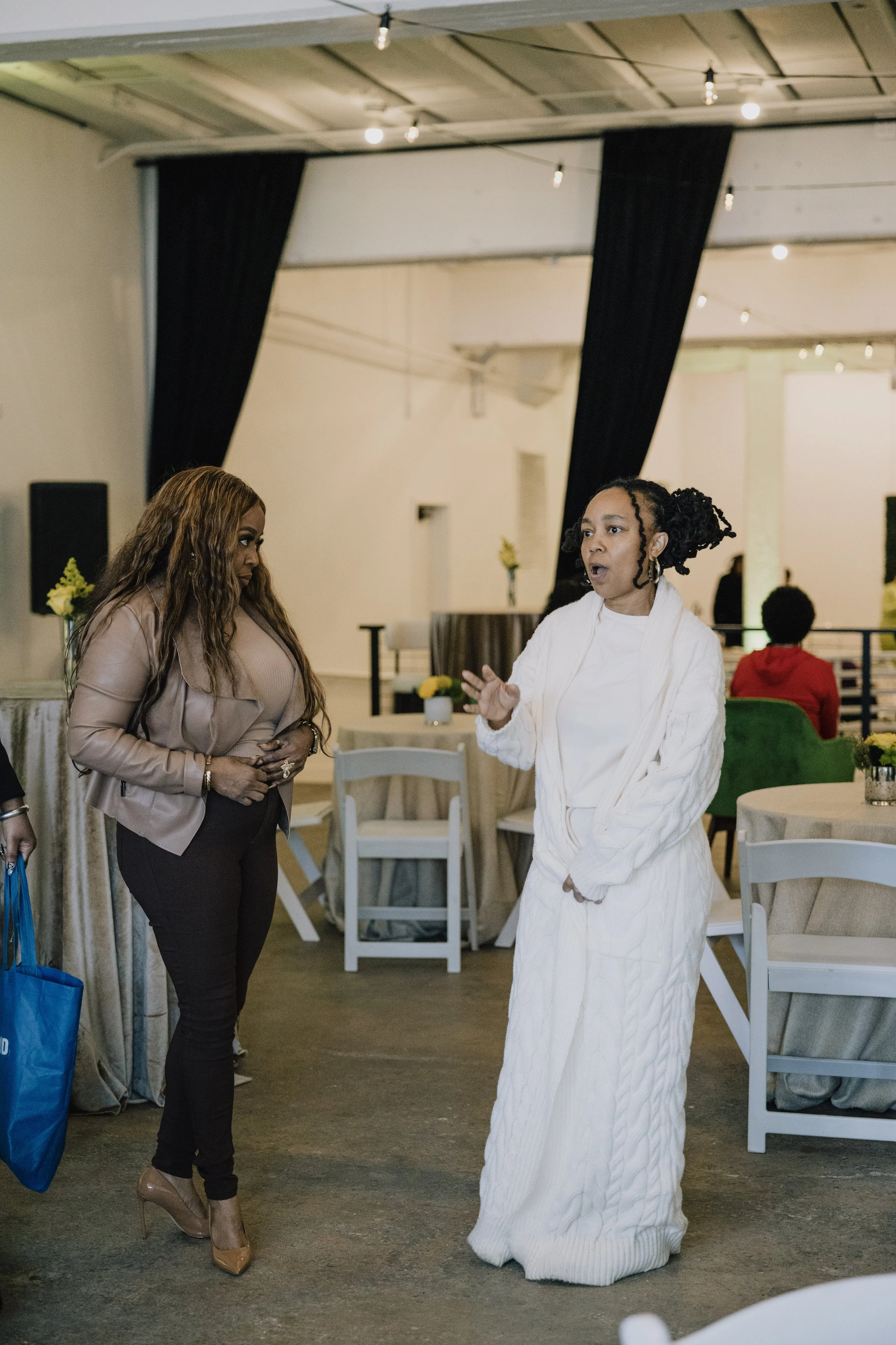 Two women having a conversation in a decorated indoor setting. One woman with dark curly hair tied up, wearing a white cardigan over a white dress, gesturing with her hand. The other woman with long wavy hair, wearing a beige jacket and black pants, holding a blue shopping bag.