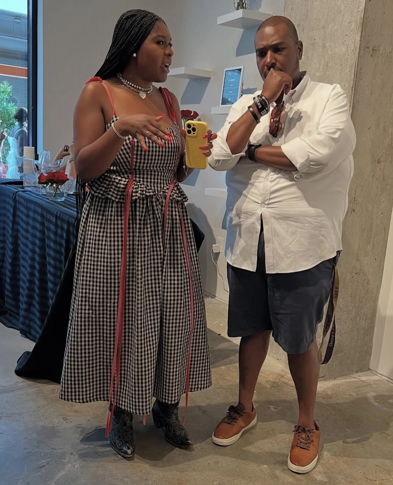 Two people standing indoors, engaged in conversation. The woman on the left is holding a phone and gesturing with her hands, wearing a checkered dress with red accents, accessories, and cowboy boots. The man on the right has his arms crossed, wearing a white shirt, dark shorts, and orange sneakers, with sunglasses hanging from his shirt.