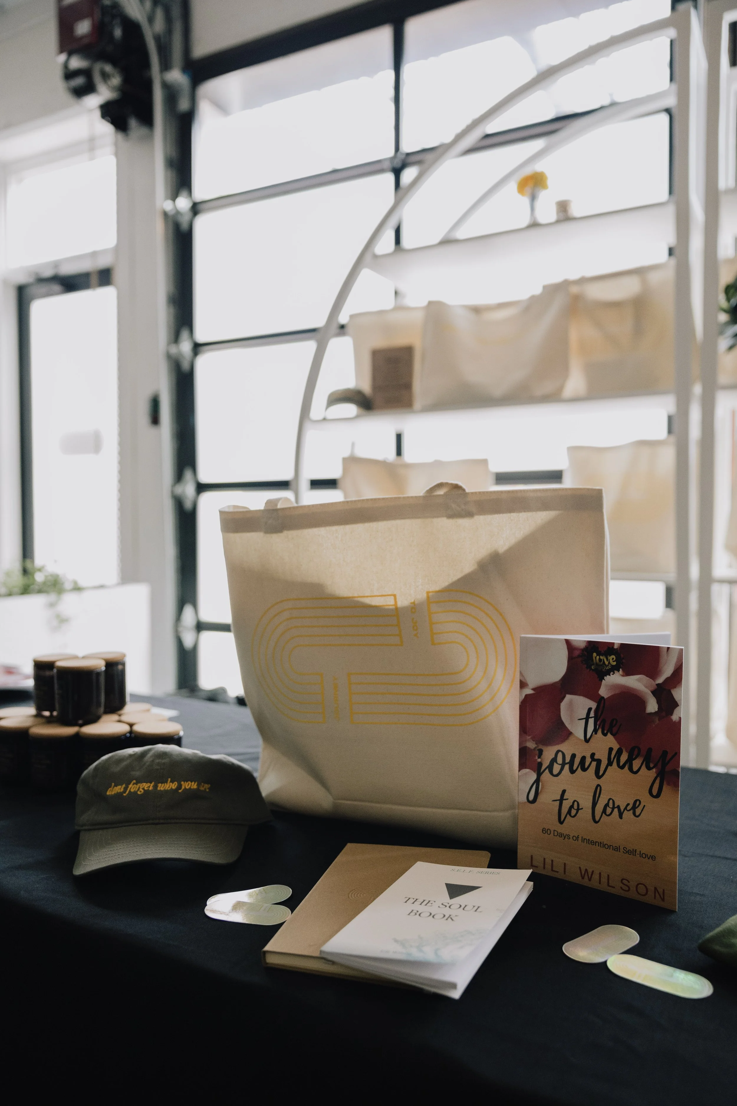Display of books, a tote bag, a baseball cap, and various items on a table at a market or event, with a window in the background.