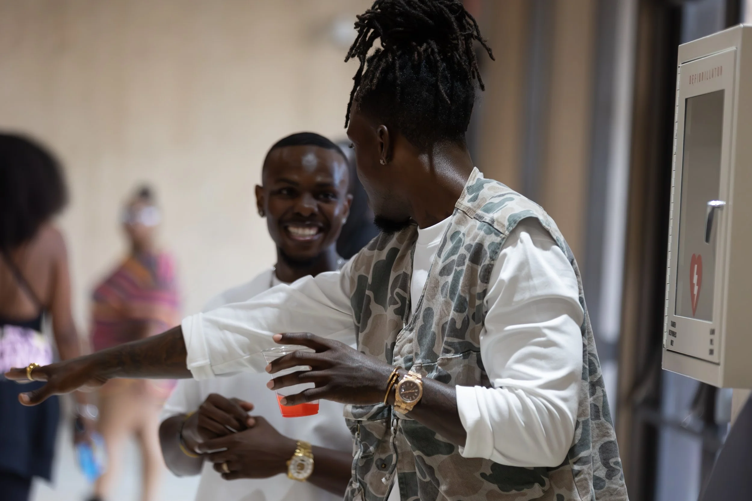 A man with dreadlocks and a camouflage vest giving a high five to a person wearing a white shirt, in a busy indoor setting.