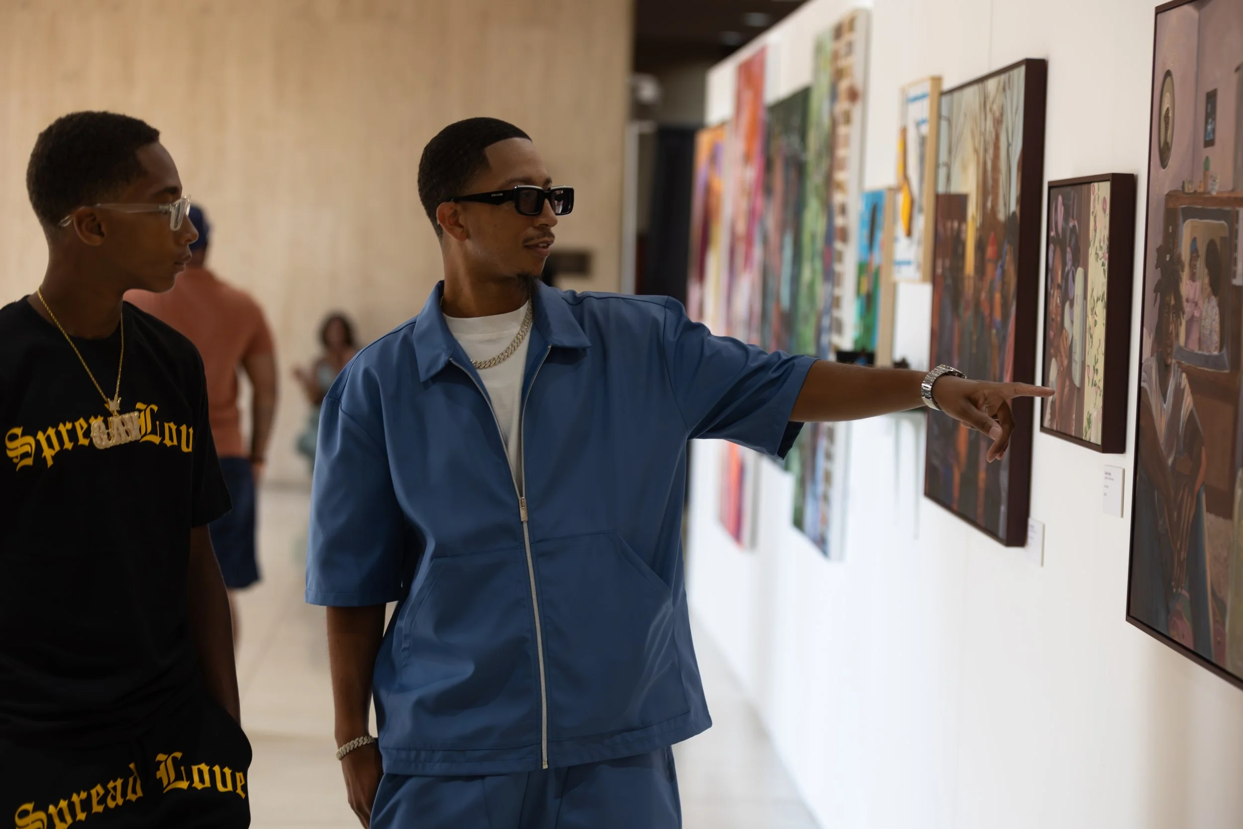 Two men are observing artwork displayed on a wall in an art gallery. One man is wearing a blue jacket and sunglasses, and the other is wearing a black T-shirt with yellow writing and glasses.
