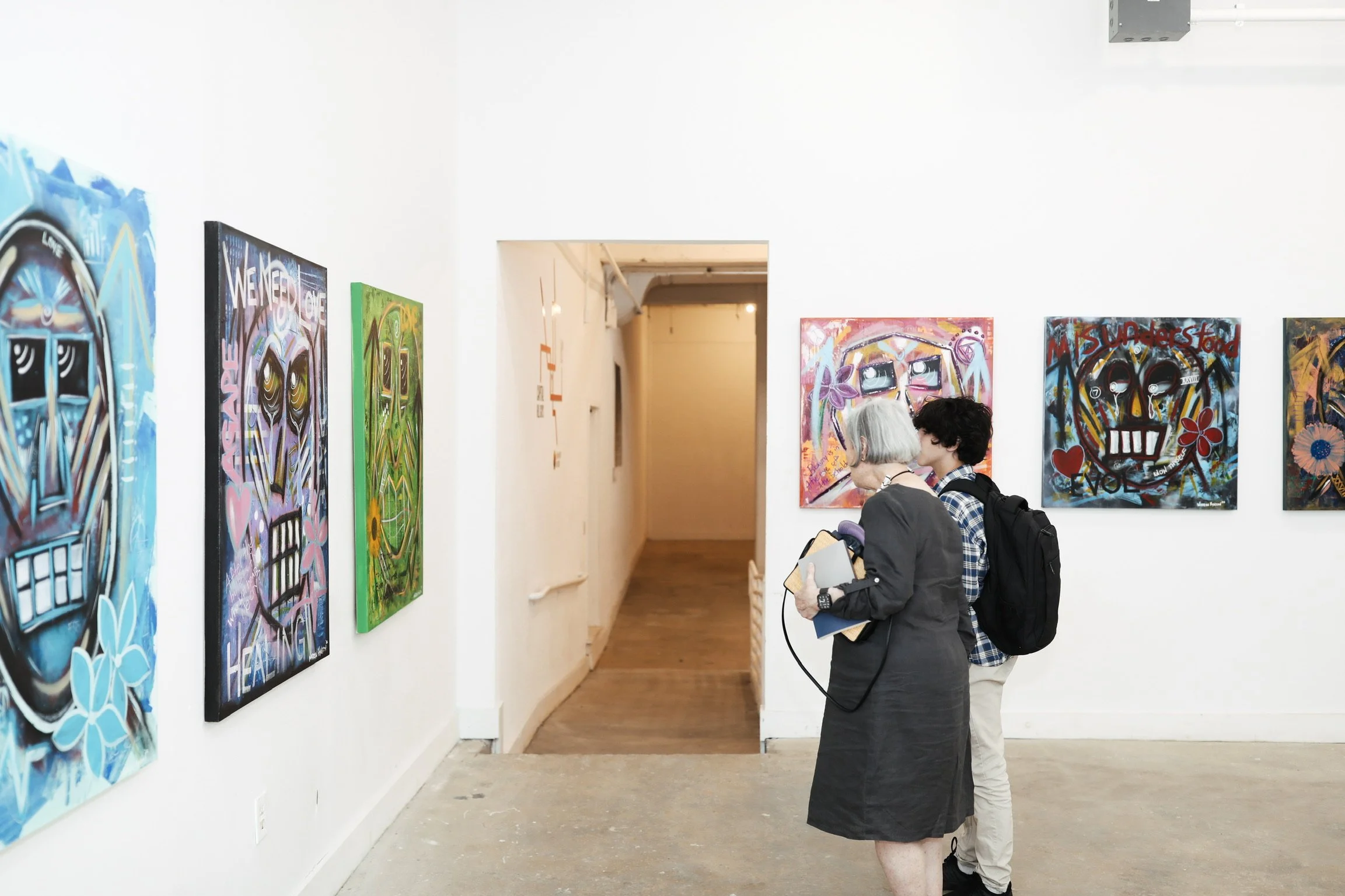 People viewing colorful paintings in an art gallery with white walls and a wooden floor.