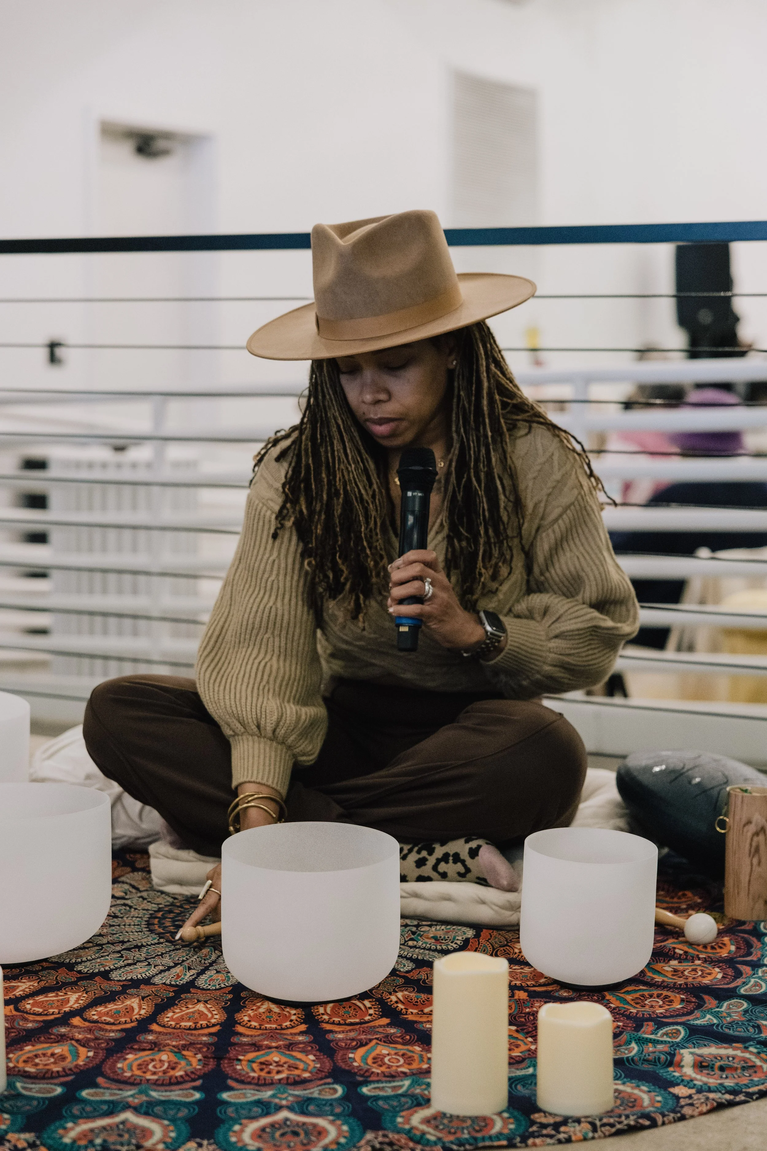 A woman with long dreadlocks and wearing a wide-brimmed tan hat sits cross-legged on a cushion, holding a microphone while surrounded by white candles on an ornate Persian-style rug, possibly in a meditation or musical setting.