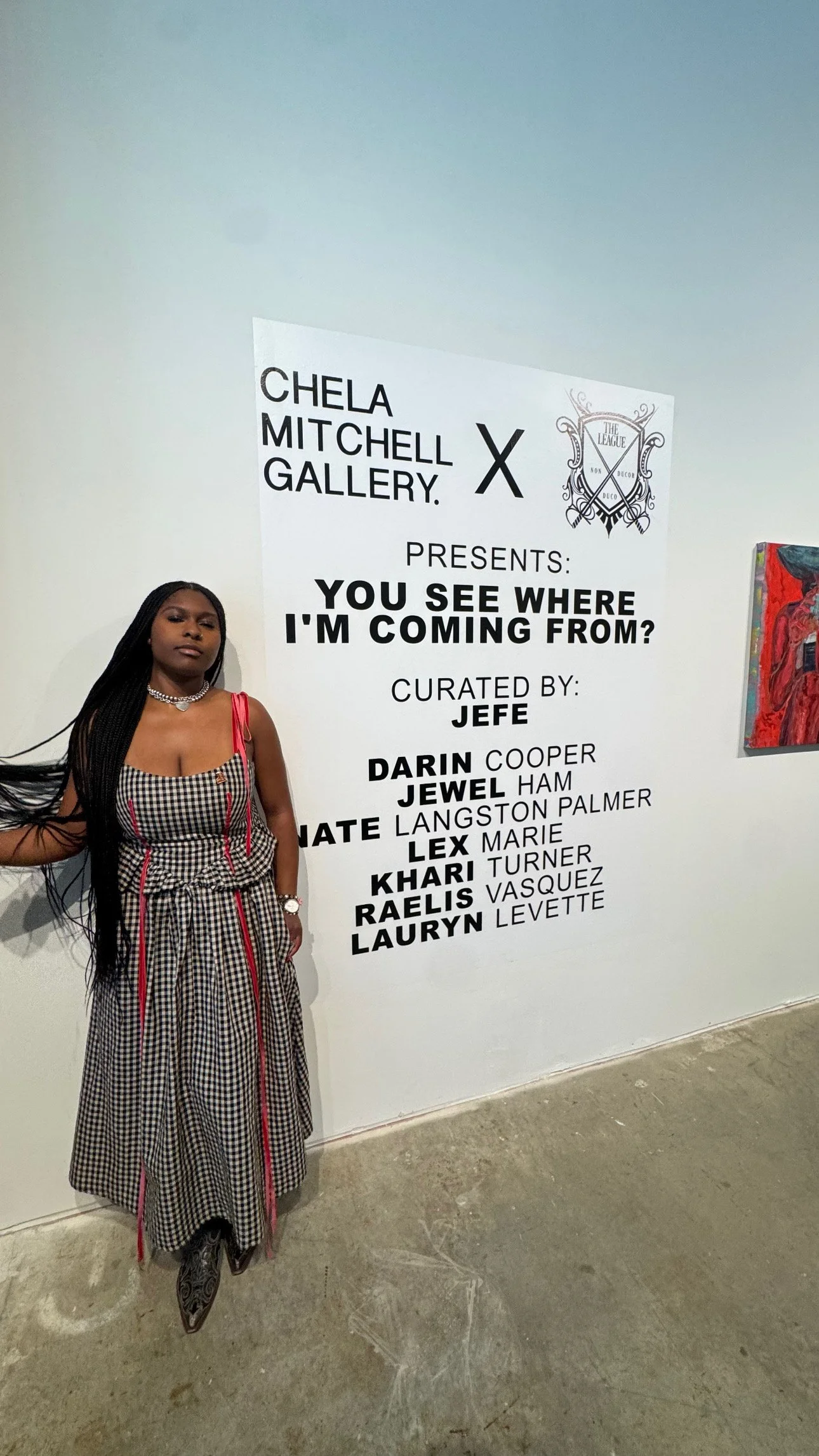A woman with long black braided hair standing next to an art exhibition poster at Chela Mitchell Gallery. The poster features the exhibition title 'You See Where I'm Coming From?' and lists various curated artists' names.