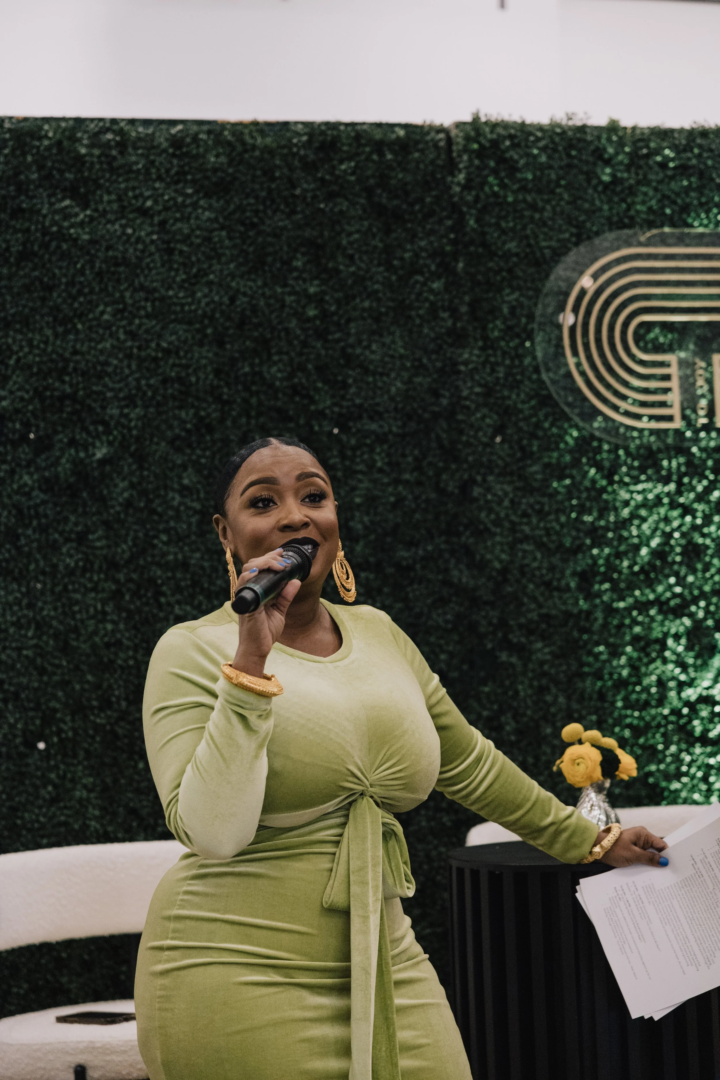 A woman in a light green dress holding a microphone and some papers, standing in front of a dark green leafy wall with a gold-colored design.