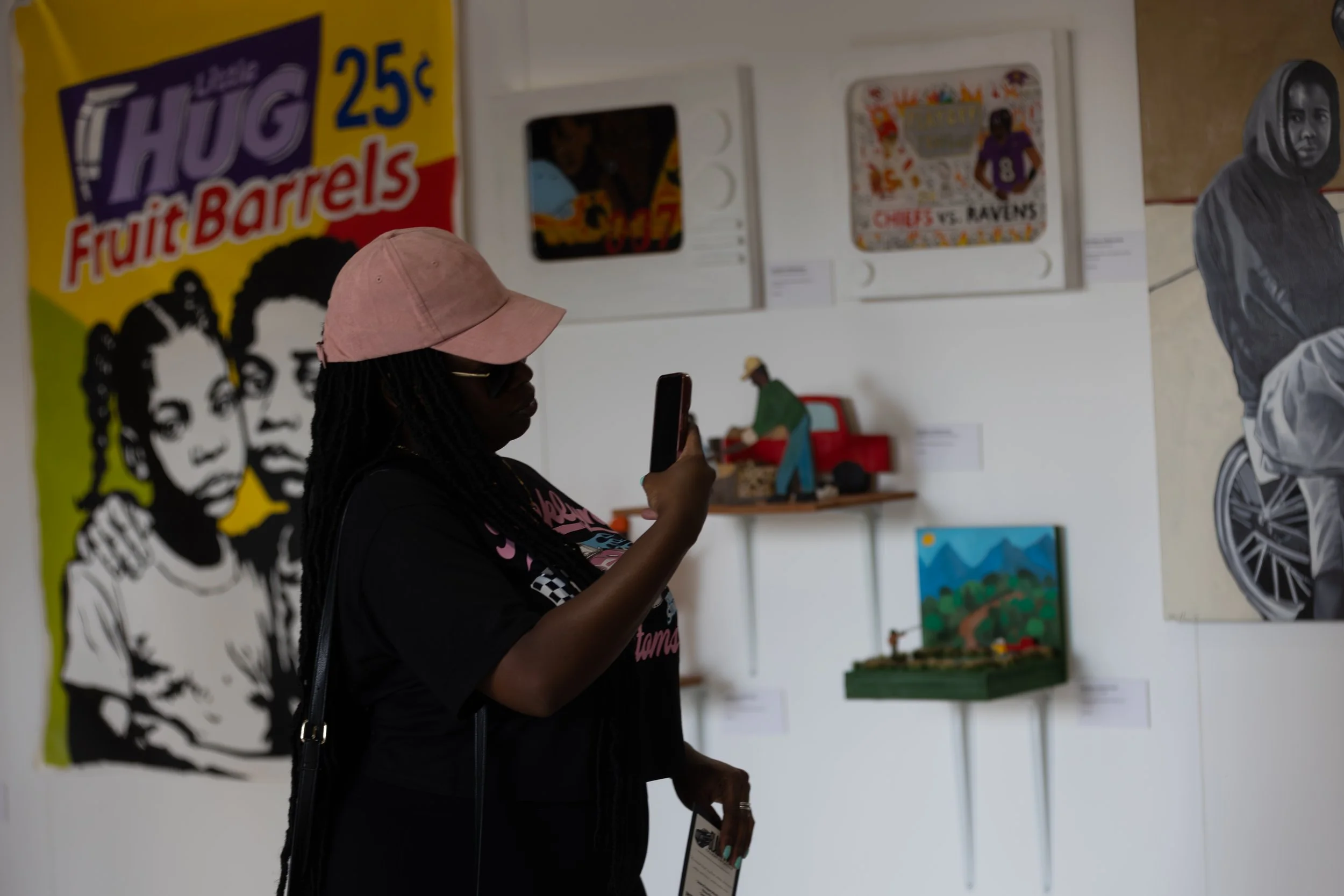 A woman wearing a pink baseball cap and black clothing is inside an art gallery, looking at her phone. The background features colorful artwork including a large poster with the text 'THUG 25¢ Fruit Barrels' and various paintings on the wall.