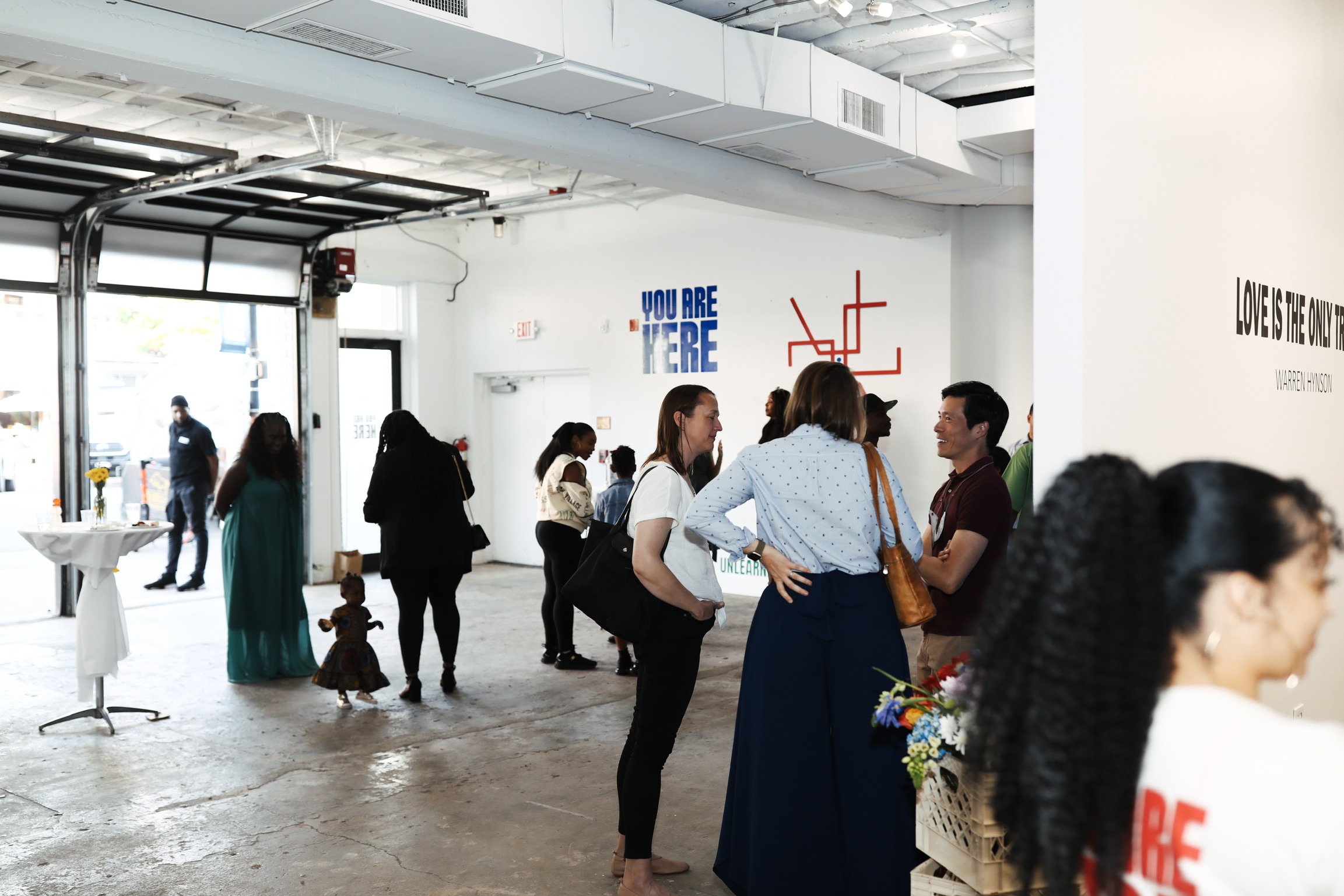 People gathered indoors at a social event, some engaged in conversations. The interior features white walls with art and motivational quotes, a high ceiling, and large garage-style doors allowing natural light to enter.