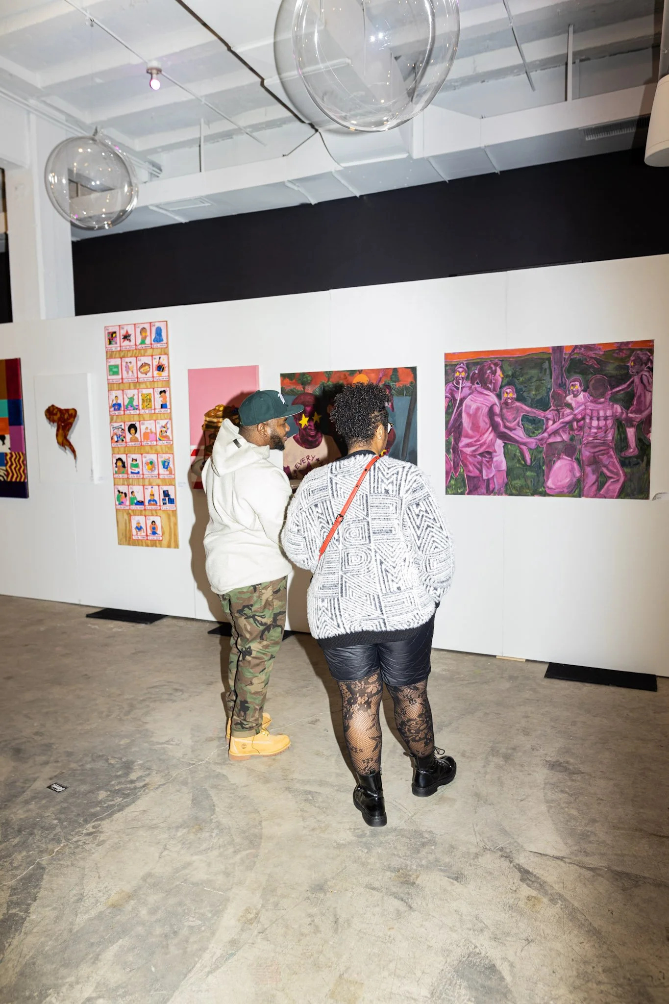 Two women and one man attending an art exhibition, observing a vibrant pink and purple painting of a group of people playing a game outdoors, as other artwork is displayed on the white walls of the gallery.