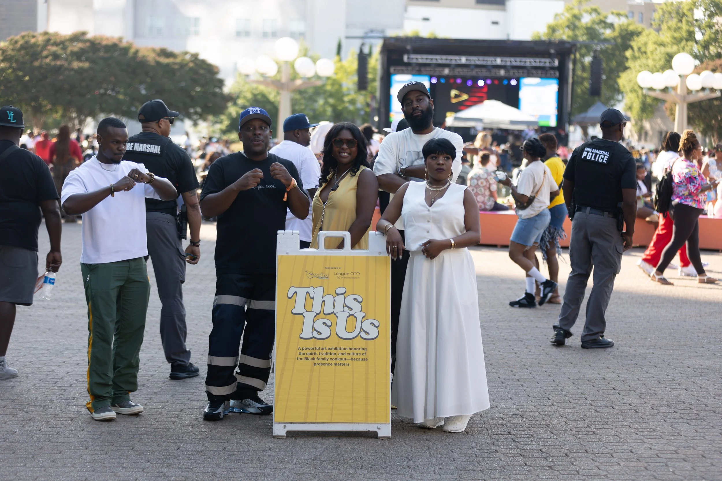 People standing outdoors at an art exhibition, including a woman in a white dress and sunglasses, surrounding a yellow sign that reads 'This Is Us' with a description of honoring Black culture and history. There is a stage with bright lights in the background, and several other visitors are visible.