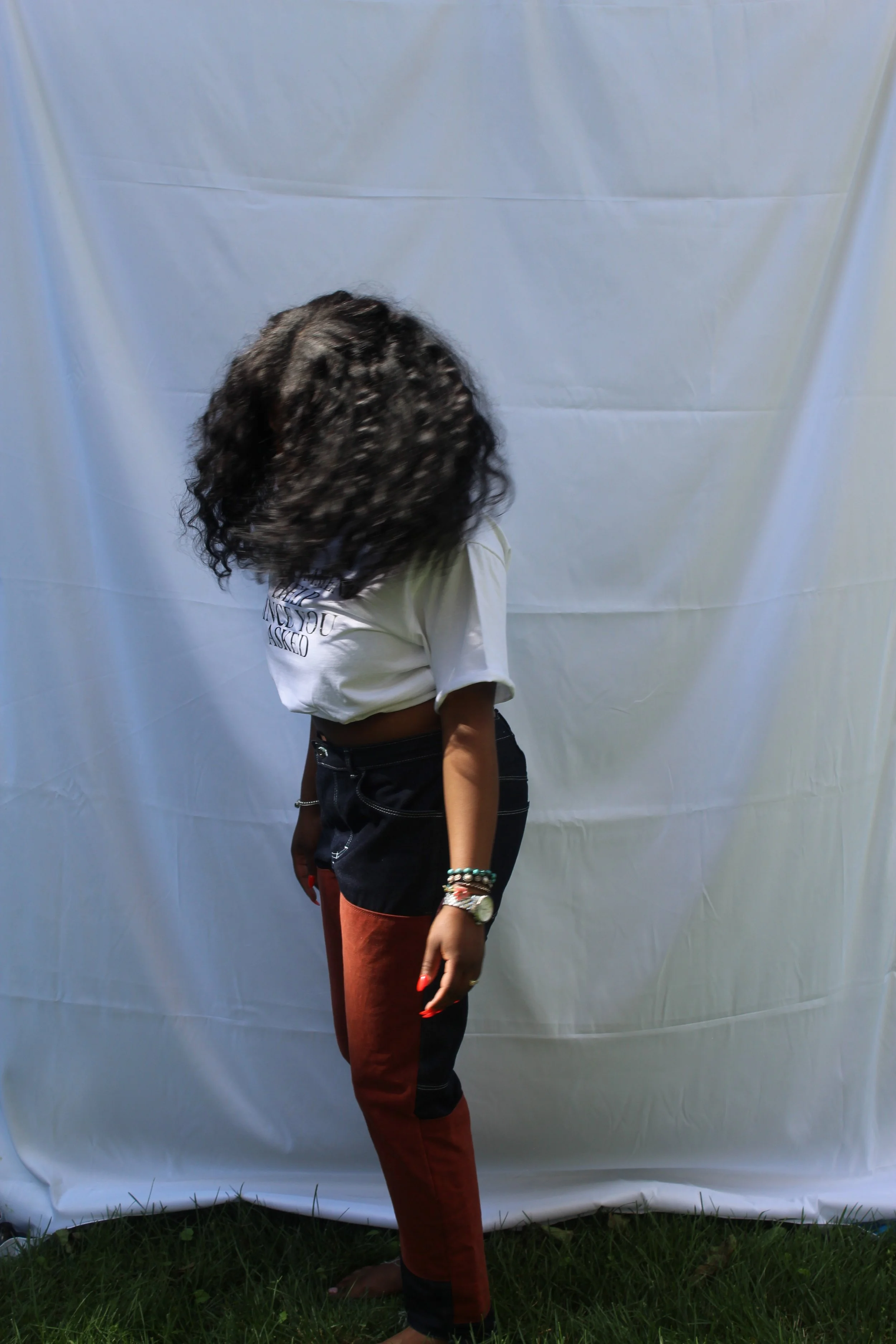 A person with curly dark hair wearing a cream T-shirt and black and red pants, standing barefoot on grass in front of a white backdrop.