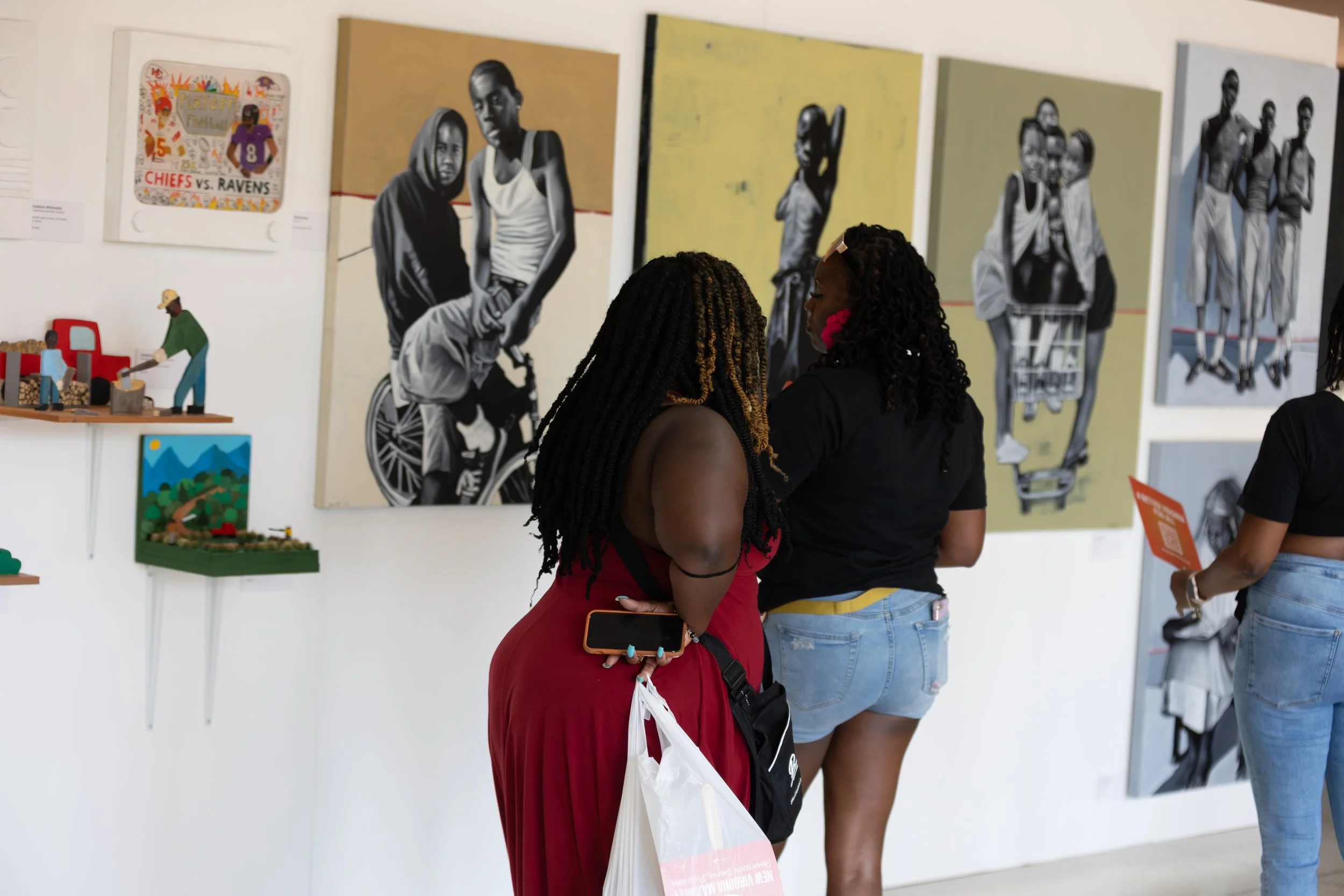 Two women with braided hair and casual clothing viewing artwork in a gallery, with one holding a phone and a bag, and other visitors visible in the background.