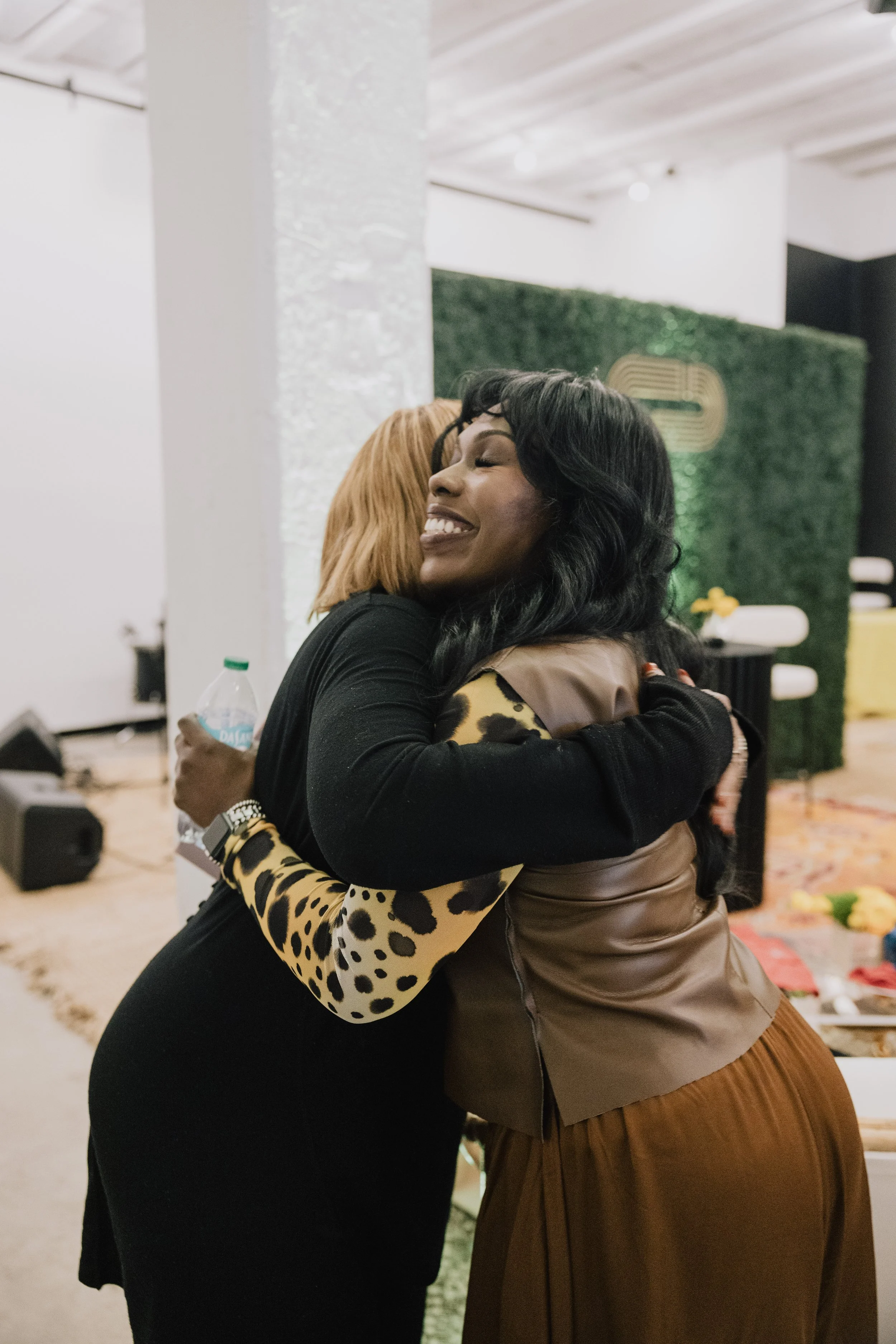 Two women hugging warmly, smiling in a warmly lit indoor setting with a green wall and decorative elements in the background.
