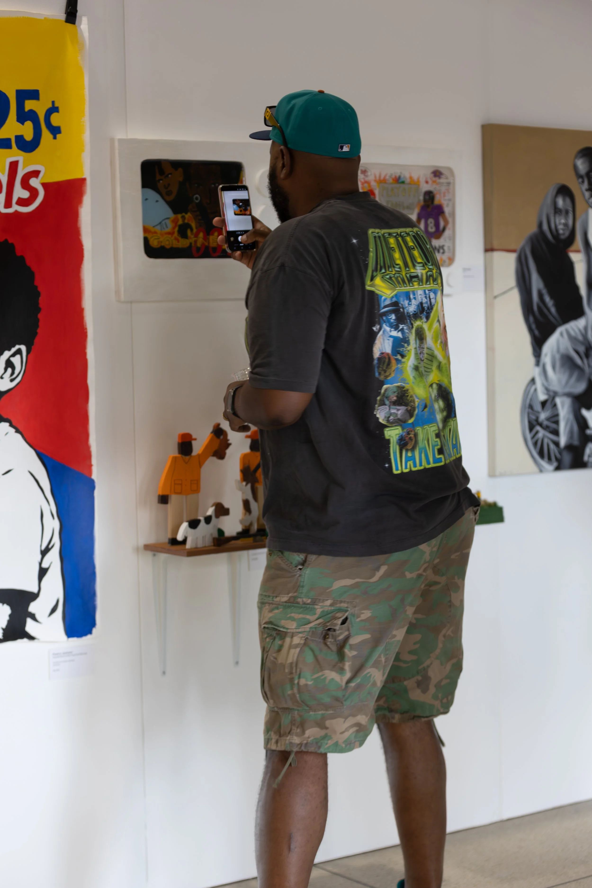A man wearing camouflage shorts, a graphic t-shirt, a teal baseball cap, and sunglasses, is taking a photo of framed artwork and wooden animal toys on a gallery wall.