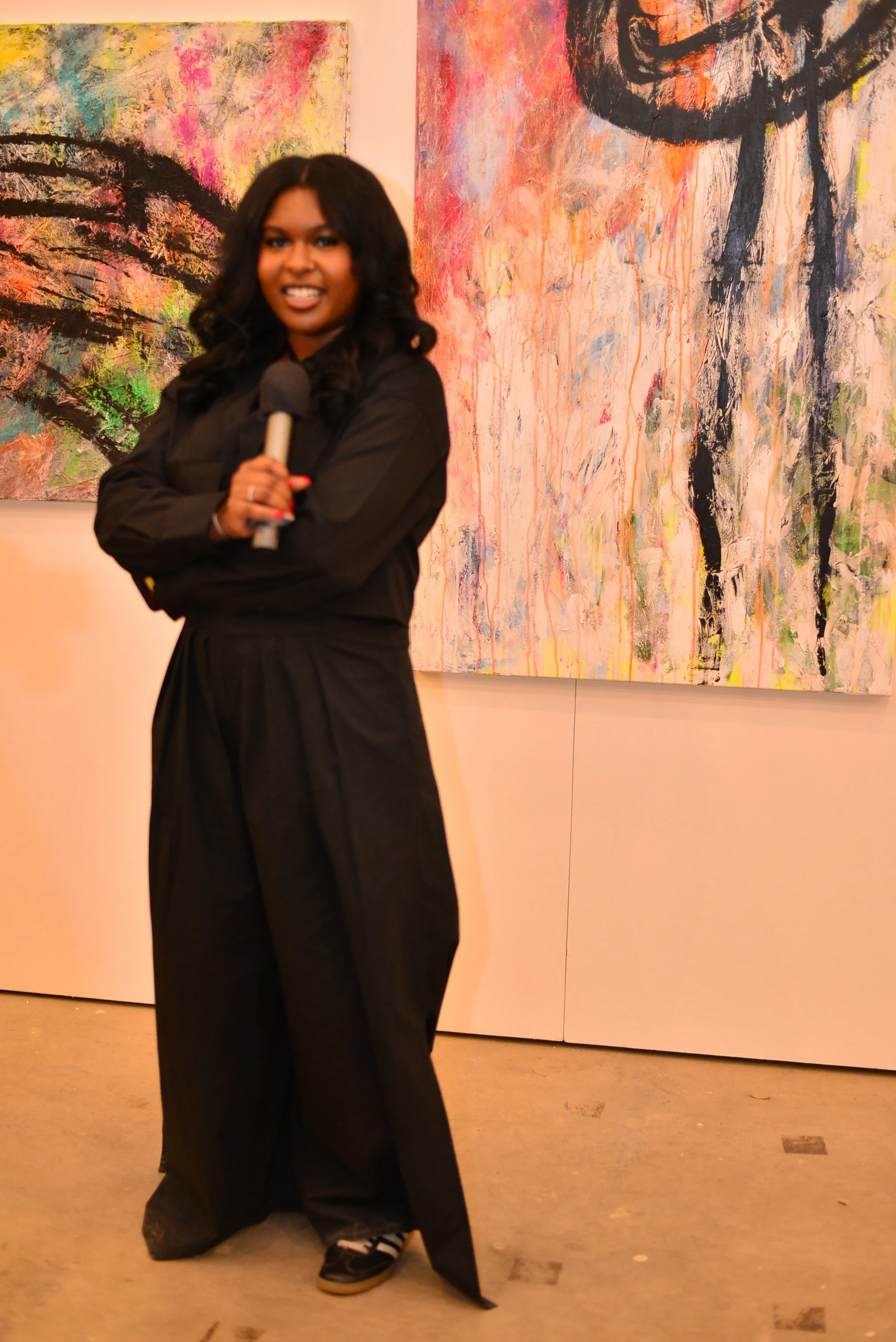 A woman with dark curly hair smiling and holding a microphone in front of abstract paintings in an art gallery.