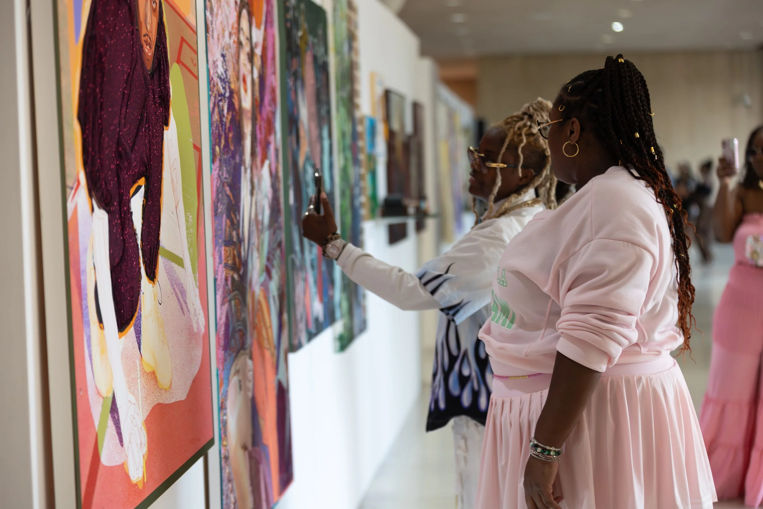 Two women with braided hair and glasses observing colorful artwork in an art gallery. One is wearing a light pink hoodie and a pink skirt, and the other is wearing a white jacket with black and white pants.