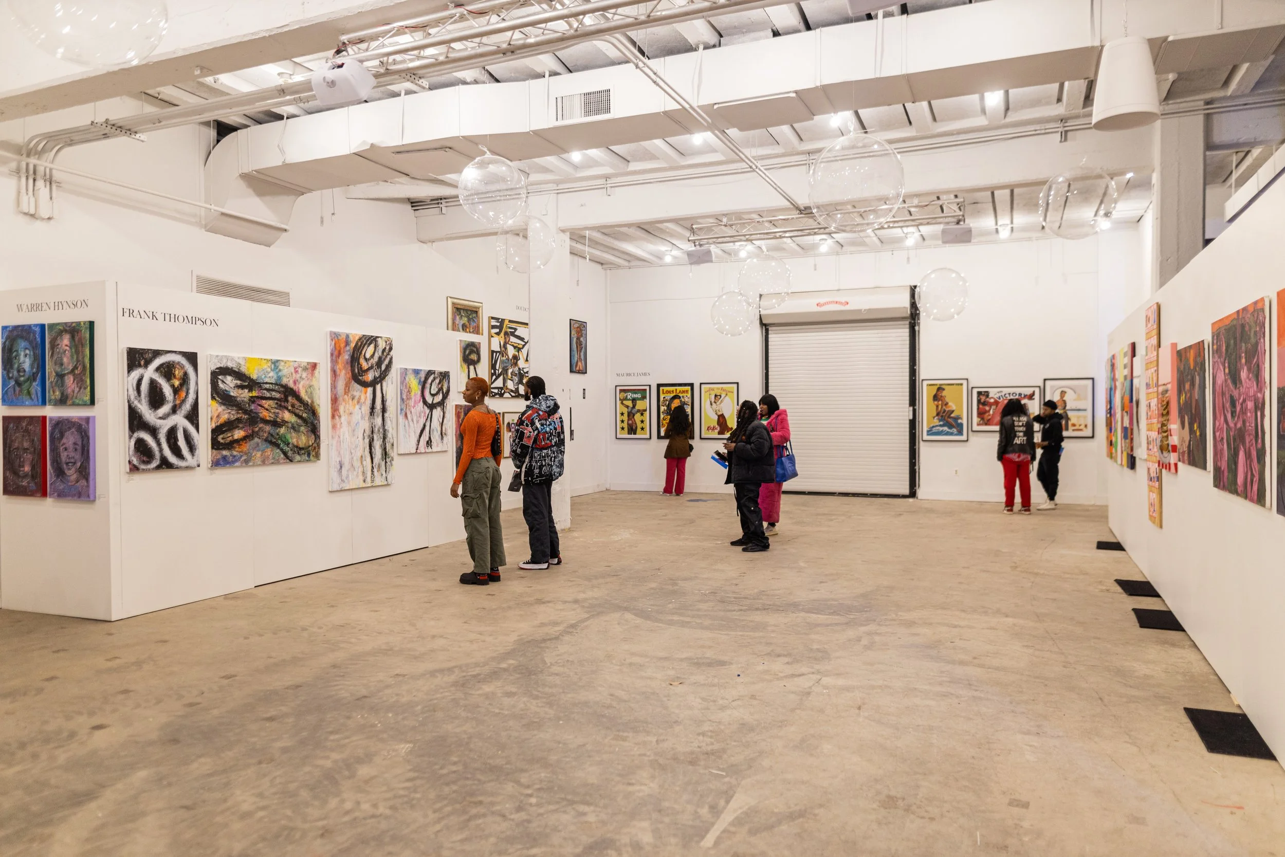 People viewing artwork in a modern art gallery with white walls, colorful paintings, and industrial ceiling with lighting fixtures.