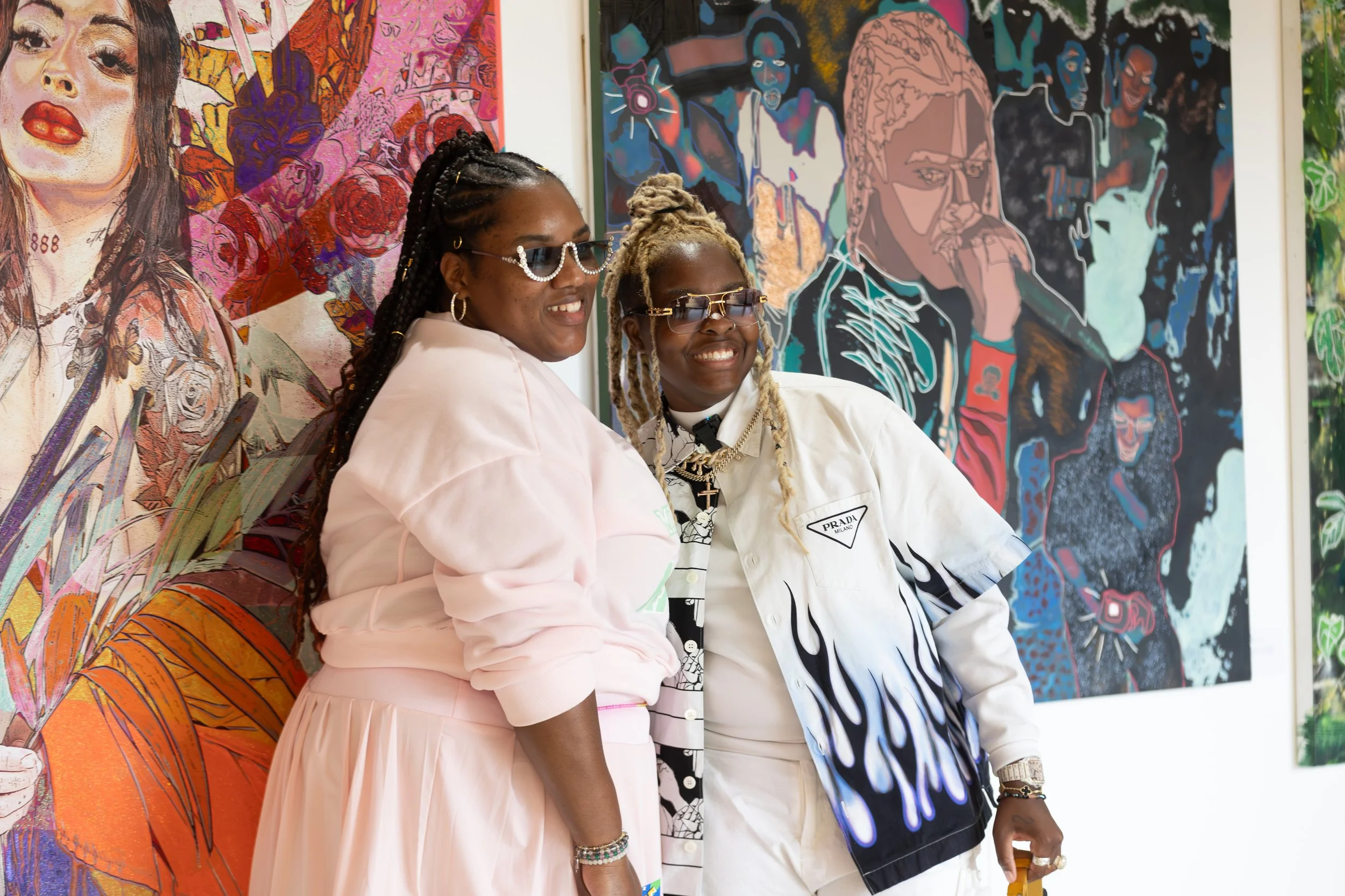 Two women smiling in front of colorful, contemporary artwork at an art gallery. One wears pink glasses and a cream jacket, the other wears dark glasses and a white Prada jacket with black and blue design.
