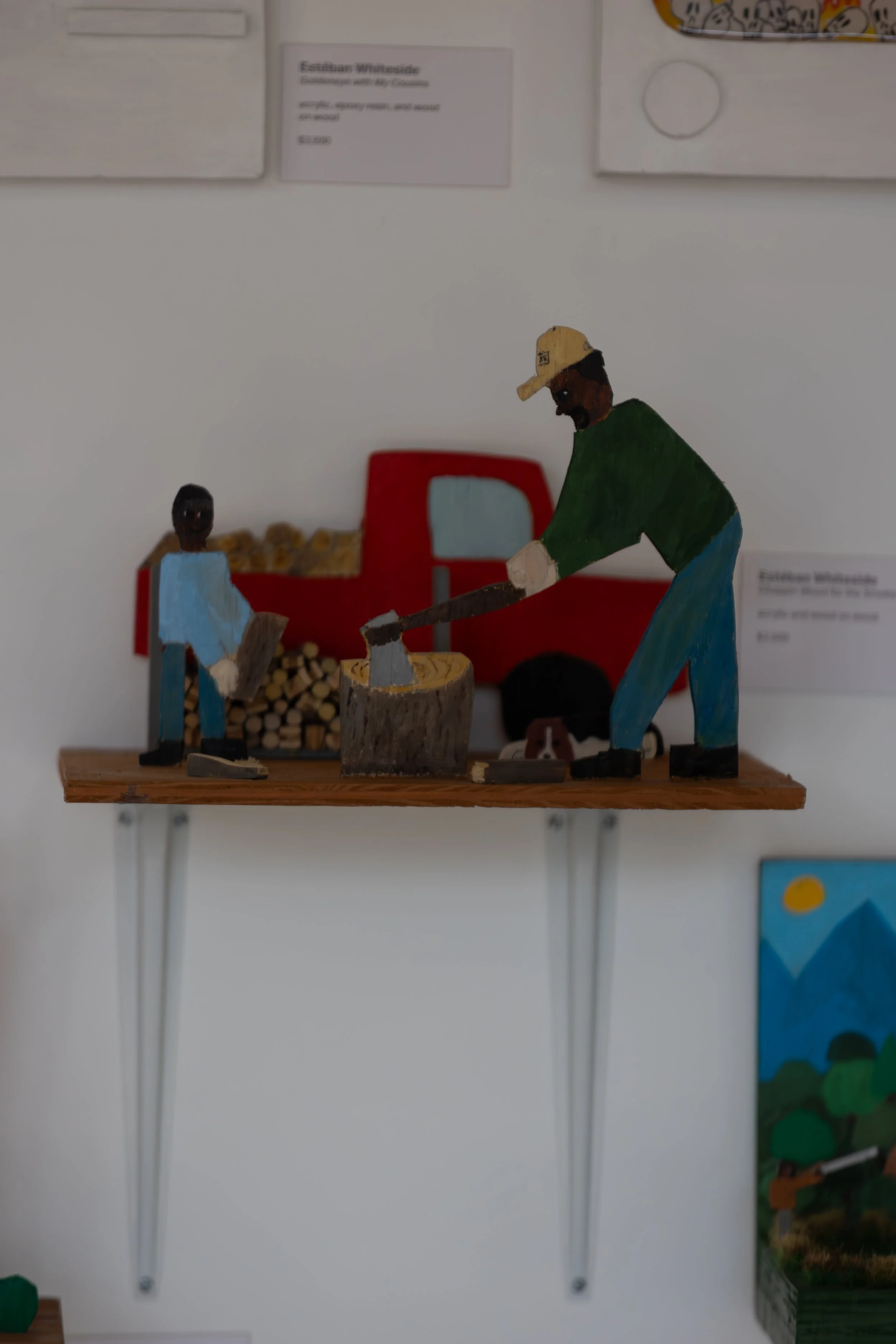 Wooden figures depicting two men working together, one with a green shirt and yellow cap, and the other in blue, with a red truck in the background filled with logs.