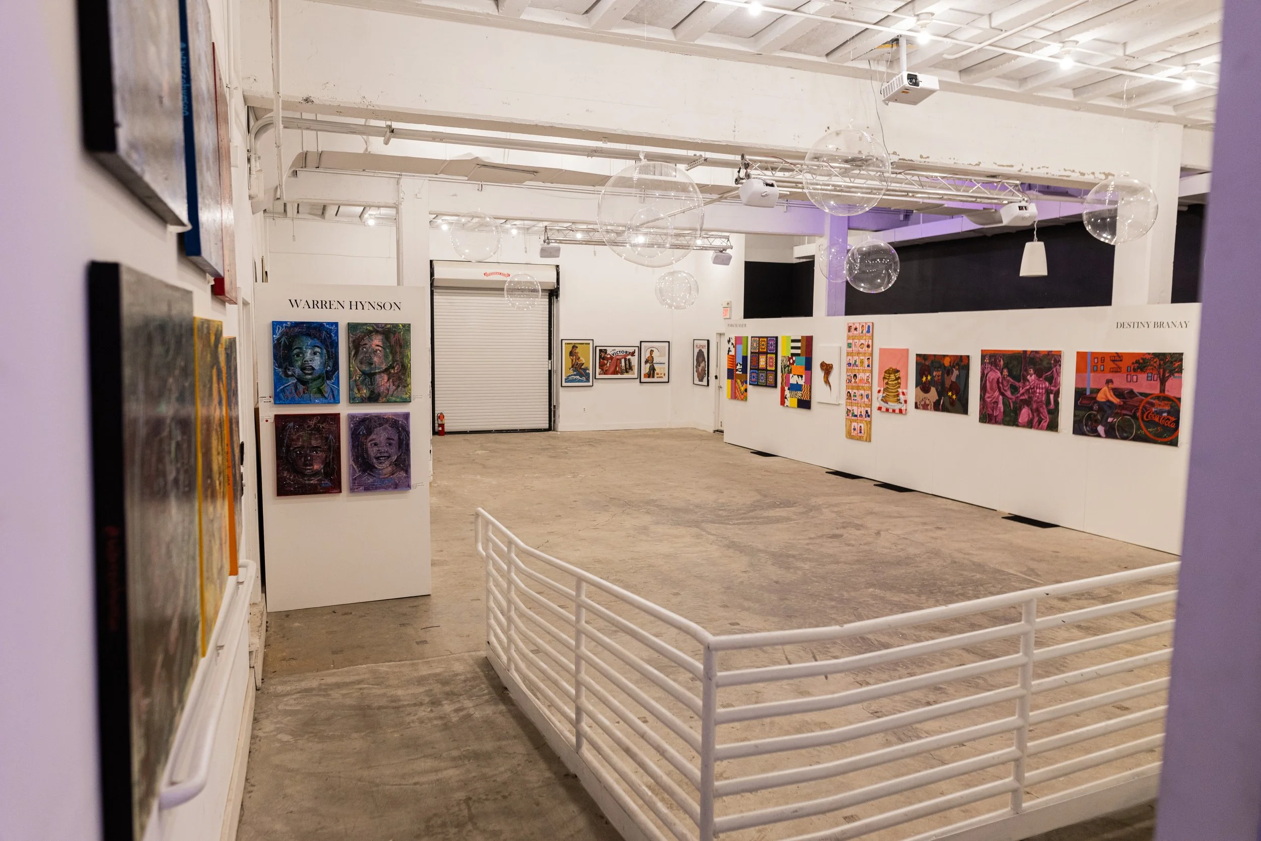 An art gallery with white walls displaying colorful paintings, some featuring portraits and abstract designs. The gallery has a wooden floor, with a white safety railing in the foreground and ceiling-mounted lighting. Artworks are arranged along the walls, with names 'WARREN HYNSON' and 'DESTINY BRANAY' visible above some of the displays.