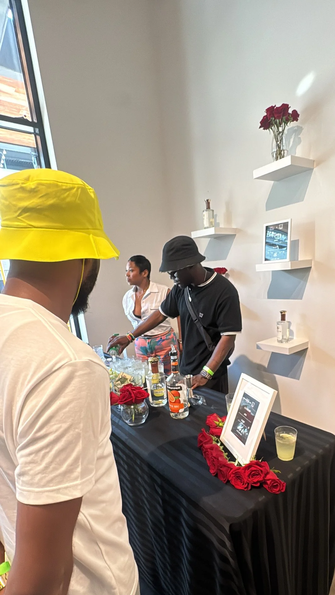 People at a bar or reception table with bottles of liquor, glasses, red roses, and a sign in a modern decorated room with white shelves and flowers.