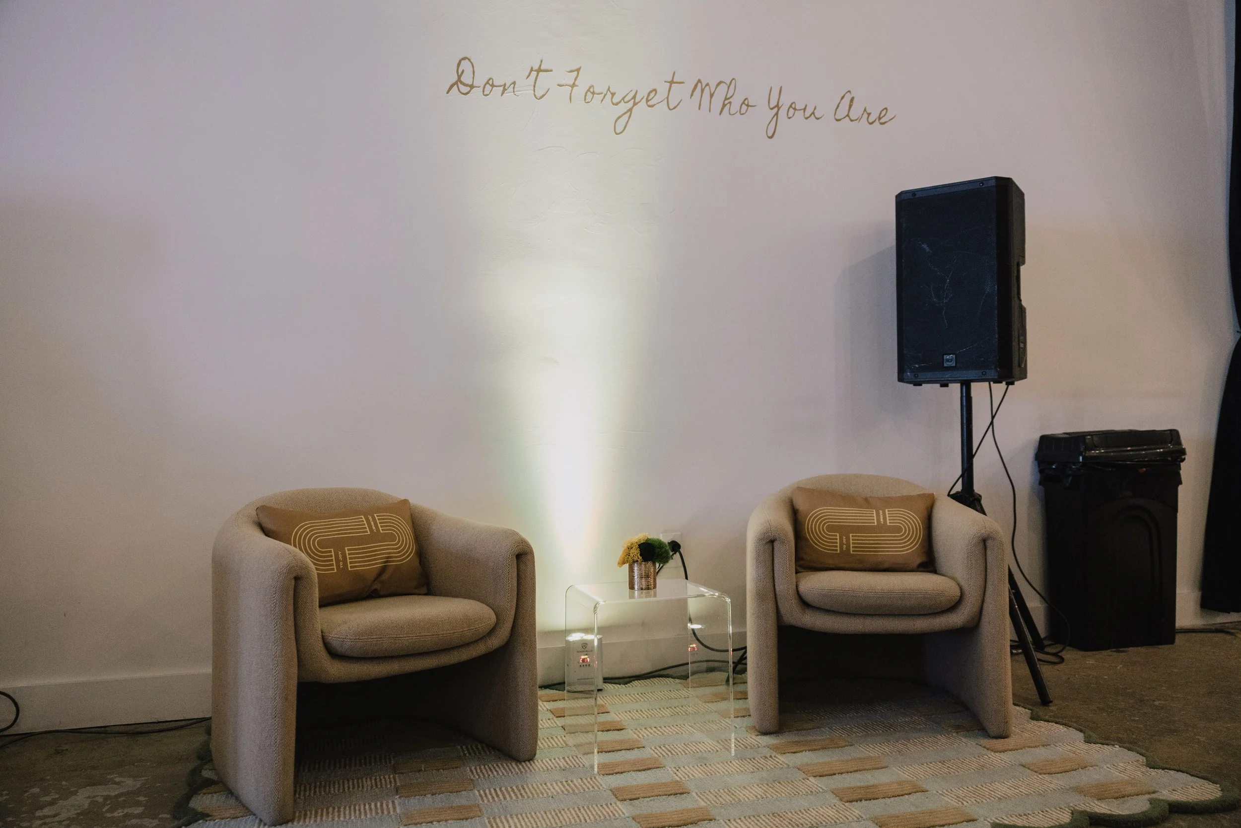 A beige sofa with a decorative pillow on each side, a small clear table with a yellow flower arrangement, a patterned rug underneath, a wall with a quote in golden cursive writing, and a large black speaker on a stand.