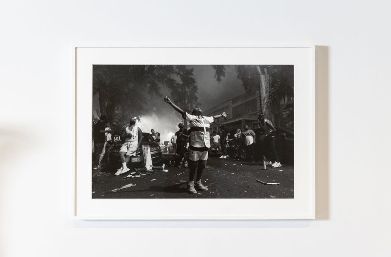 Black and white photo of a street scene with people, some appear to be running or involved in chaos, and there is smoke or fire in the background.
