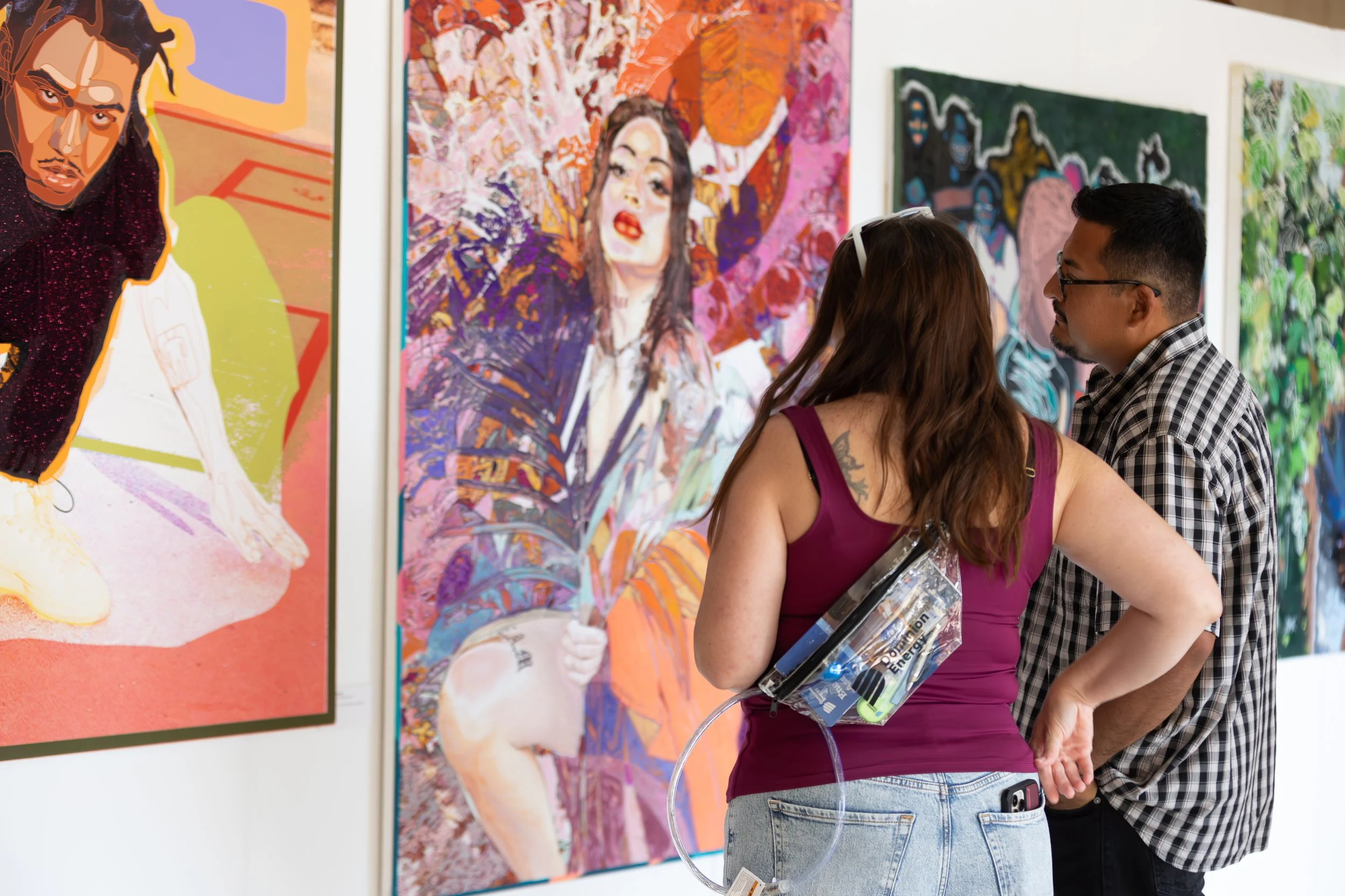 Two visitors, a woman with long brown hair and a man with short dark hair and glasses, are looking at colorful abstract portraits in an art gallery.