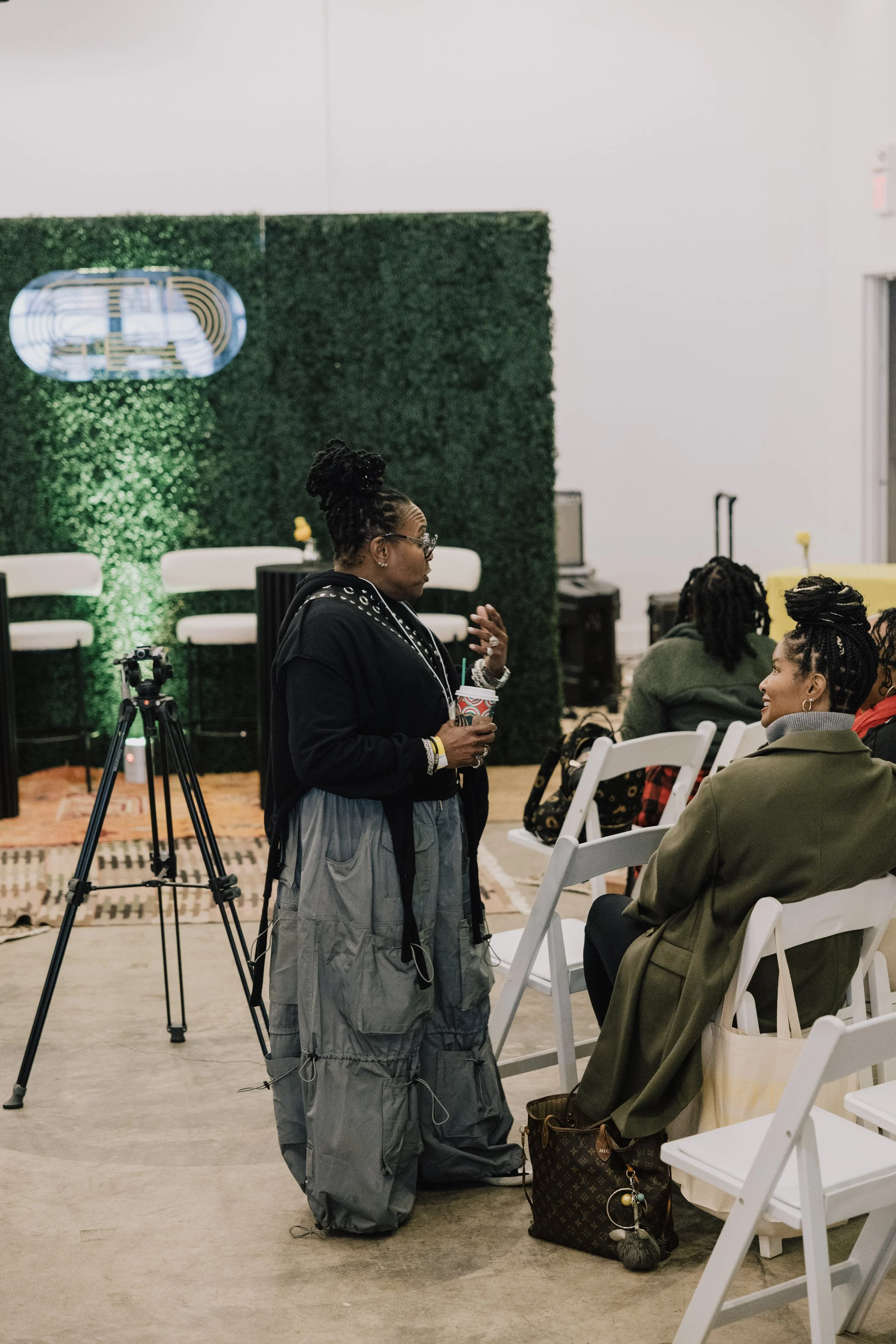A woman with glasses and a bun hairdo, dressed in a black hoodie and gray pants, holding a cup, and talking to a seated woman with braided hair in a gray jacket at an indoor event. There is a green wall with a logo in the background, and chairs and bags are visible.
