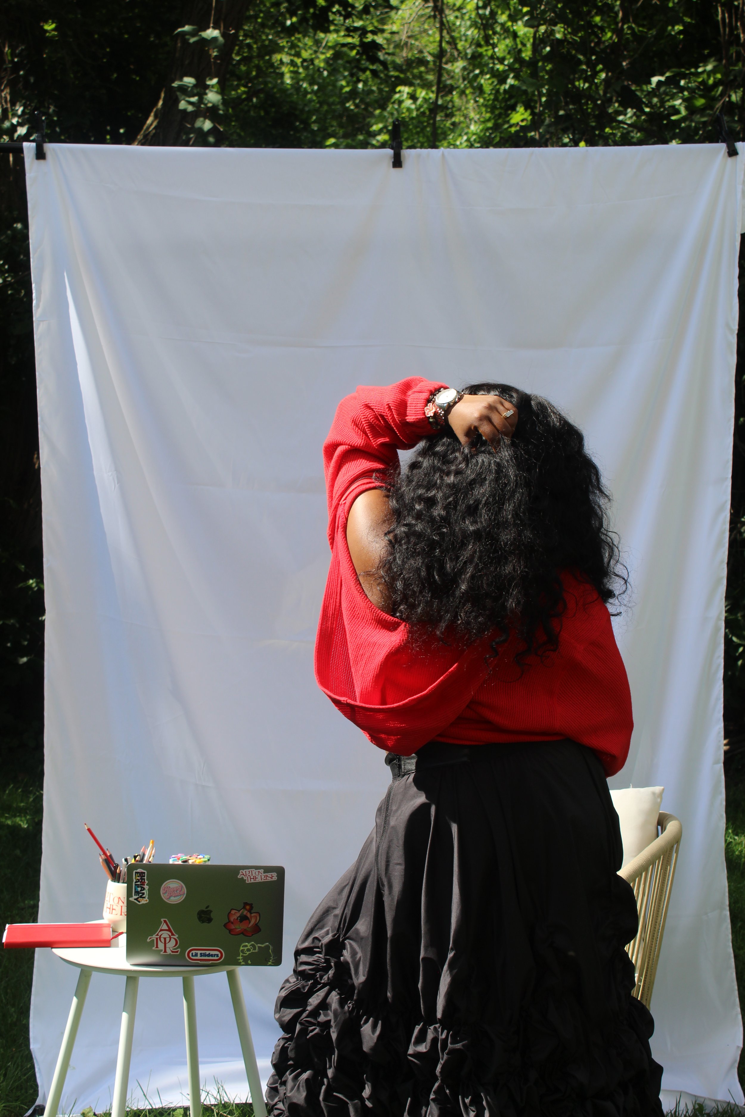 A woman with curly hair wearing a red sweater and black skirt sits in front of a white curtain set outdoors. A small white table with a laptop, stationery, and stickers is in front of her, and various supplies are on the ground. Trees are visible behind the curtain.