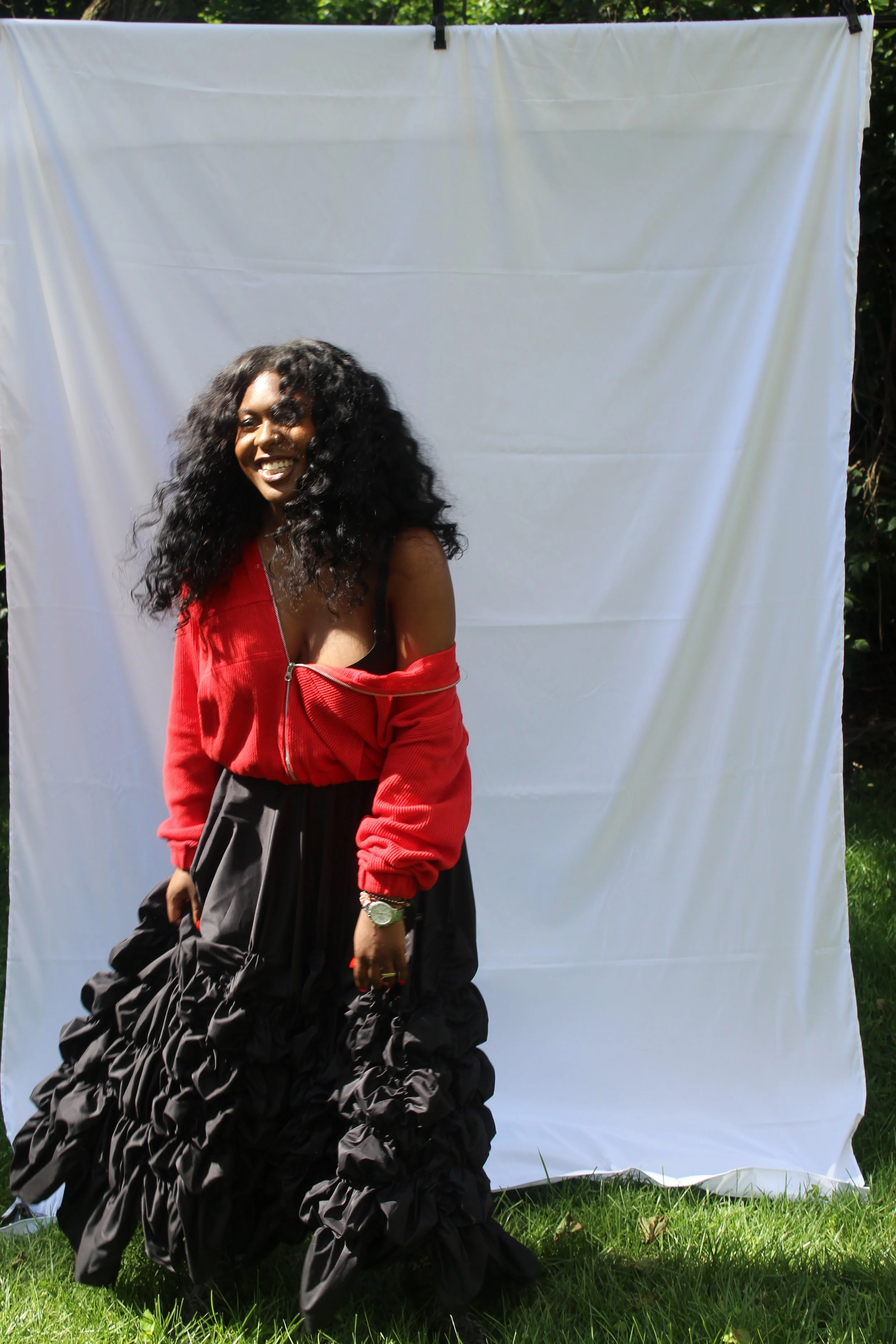 Woman standing outdoors in front of a white fabric backdrop, wearing a red top and black voluminous skirt, smiling, with lush greenery visible behind the backdrop.