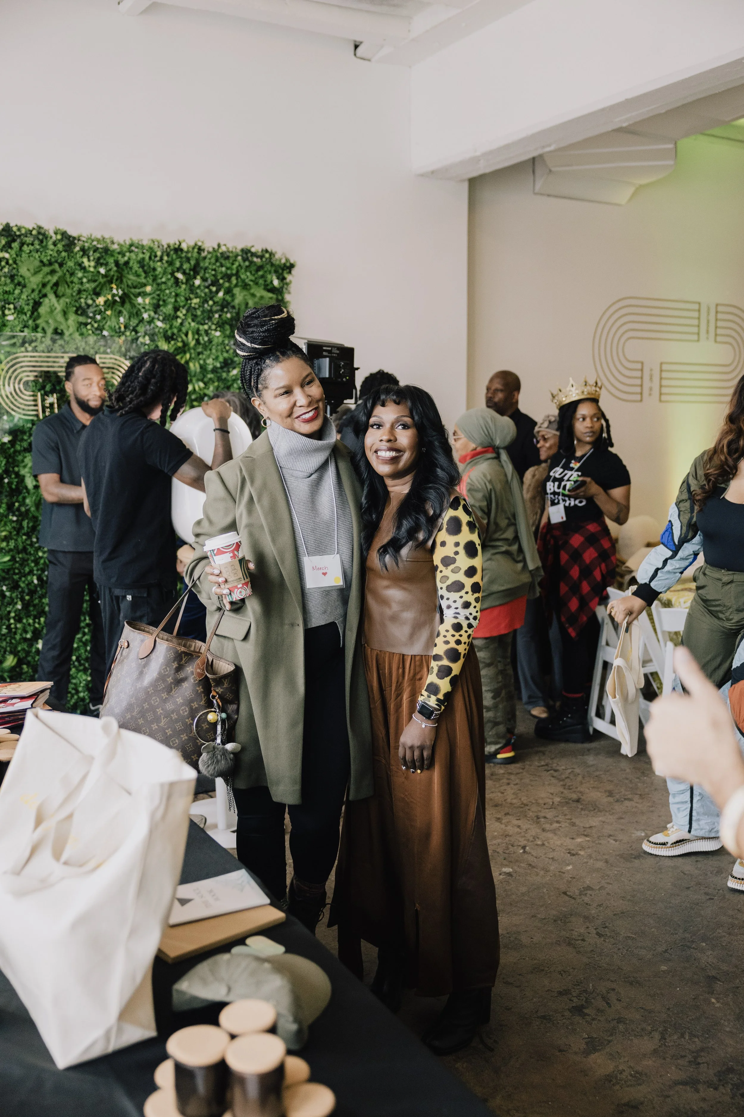Two women standing together at an indoor event, both smiling at the camera. One woman is holding a cup of coffee, and there are people in the background, some wearing crowns. The setting appears to be casual with tables, a green wall, and some decorative elements.