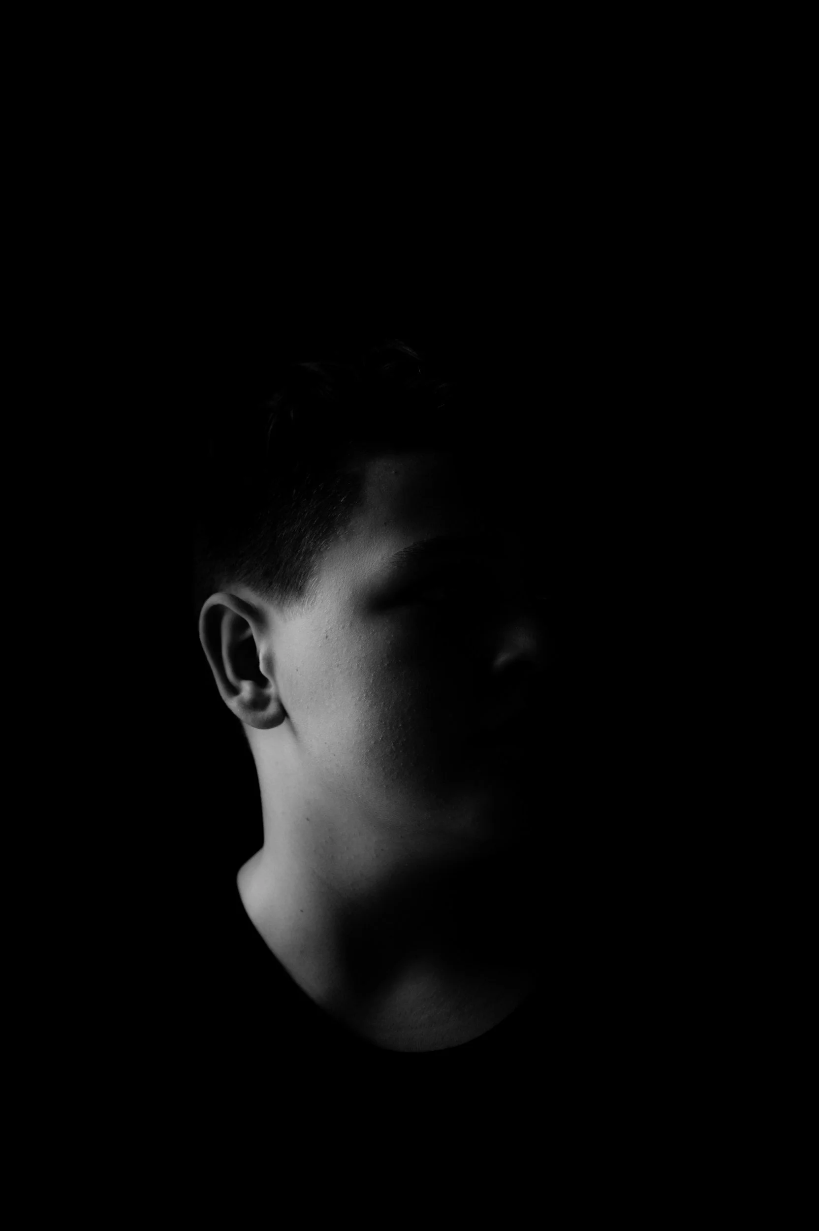 A close-up black and white photo of a person's face, showing the ear and side of the face with short hair, against a black background.