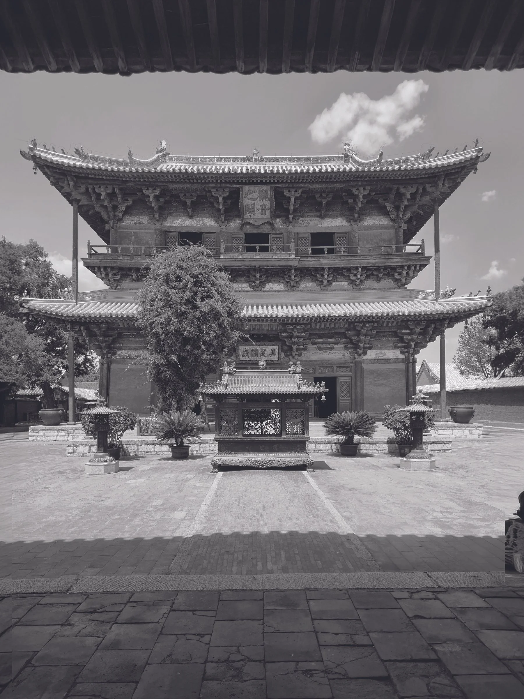 A traditional Chinese pagoda-style building with ornate roofing and decorative features, surrounded by potted plants and trees, under a partly cloudy sky.