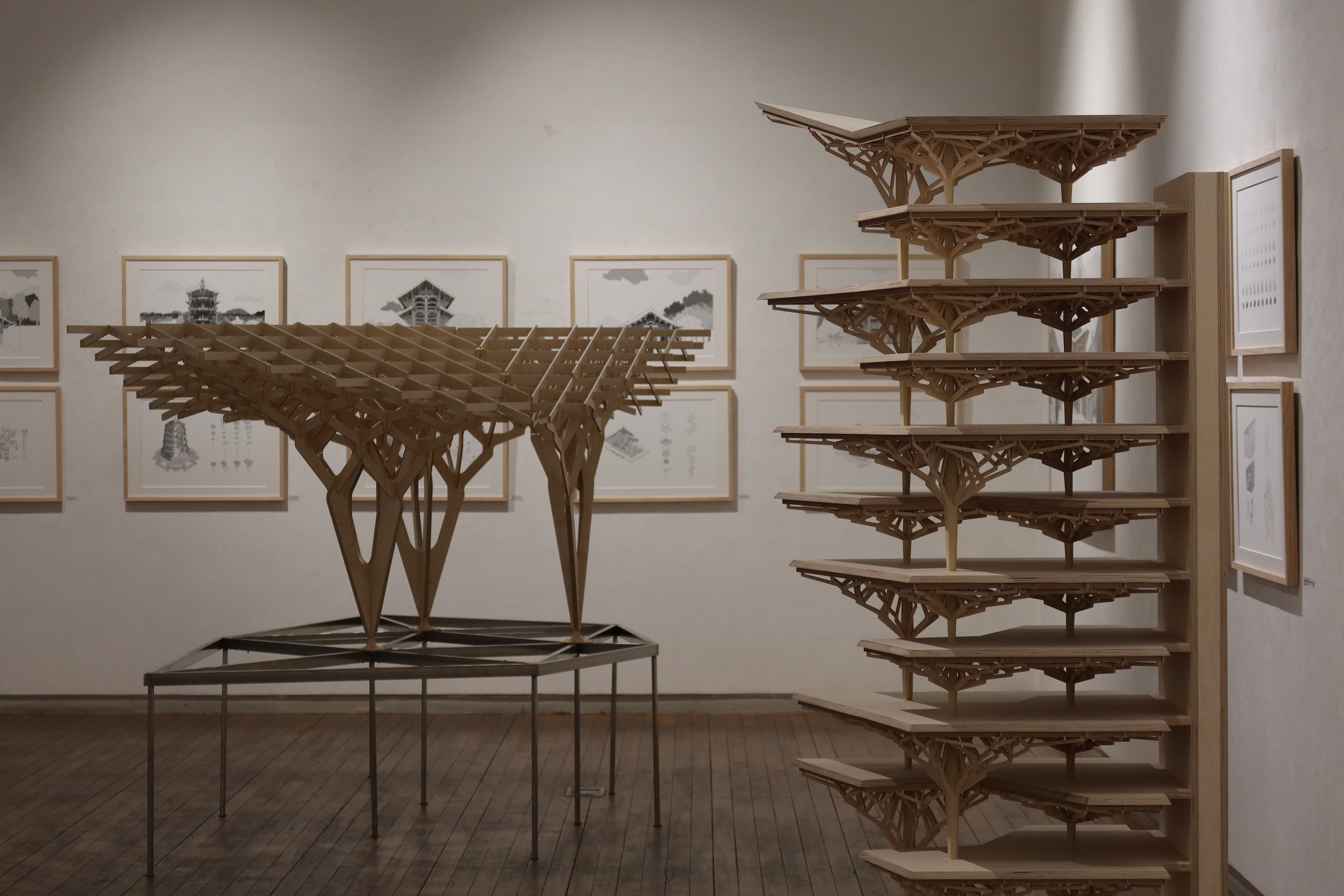 Wooden architectural models resembling trees and structures are displayed in an art gallery, with framed black-and-white architectural drawings on the wall behind.