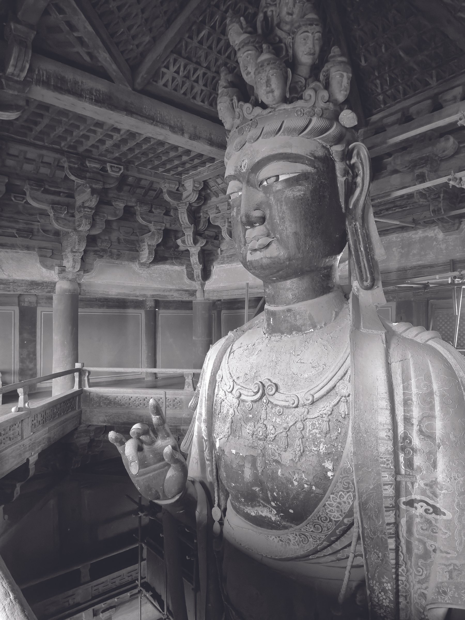 A large ancient sculpture of a female deity or figure with intricate carvings on her clothing and ornaments, displayed indoors with wooden beams and decorative architecture in the background.
