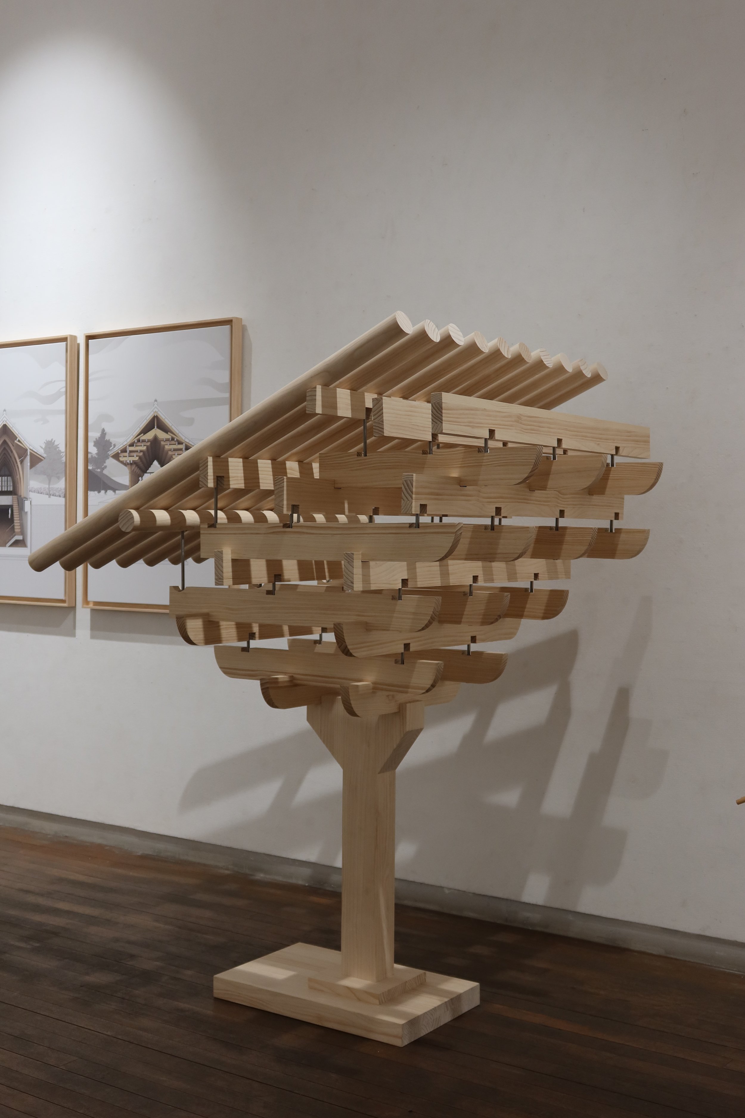 A wooden sculpture resembling a stylized tree or plant, displayed in an art gallery with framed artworks in the background.