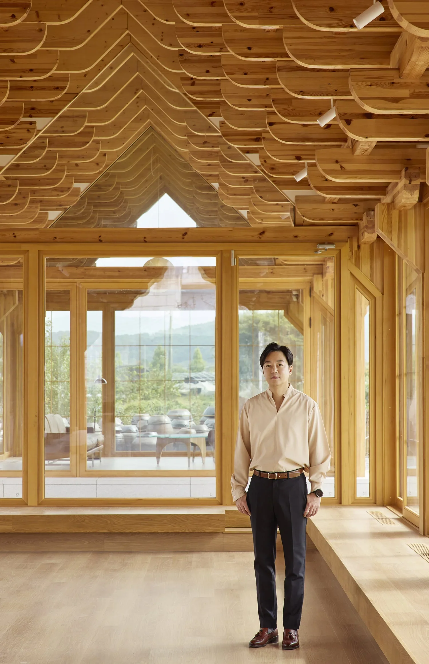 A man dressed in a beige shirt and dark pants stands inside a wooden building with large windows and a high, curved wooden ceiling. The view through the windows shows a parking lot with several cars and a landscape of trees and hills.