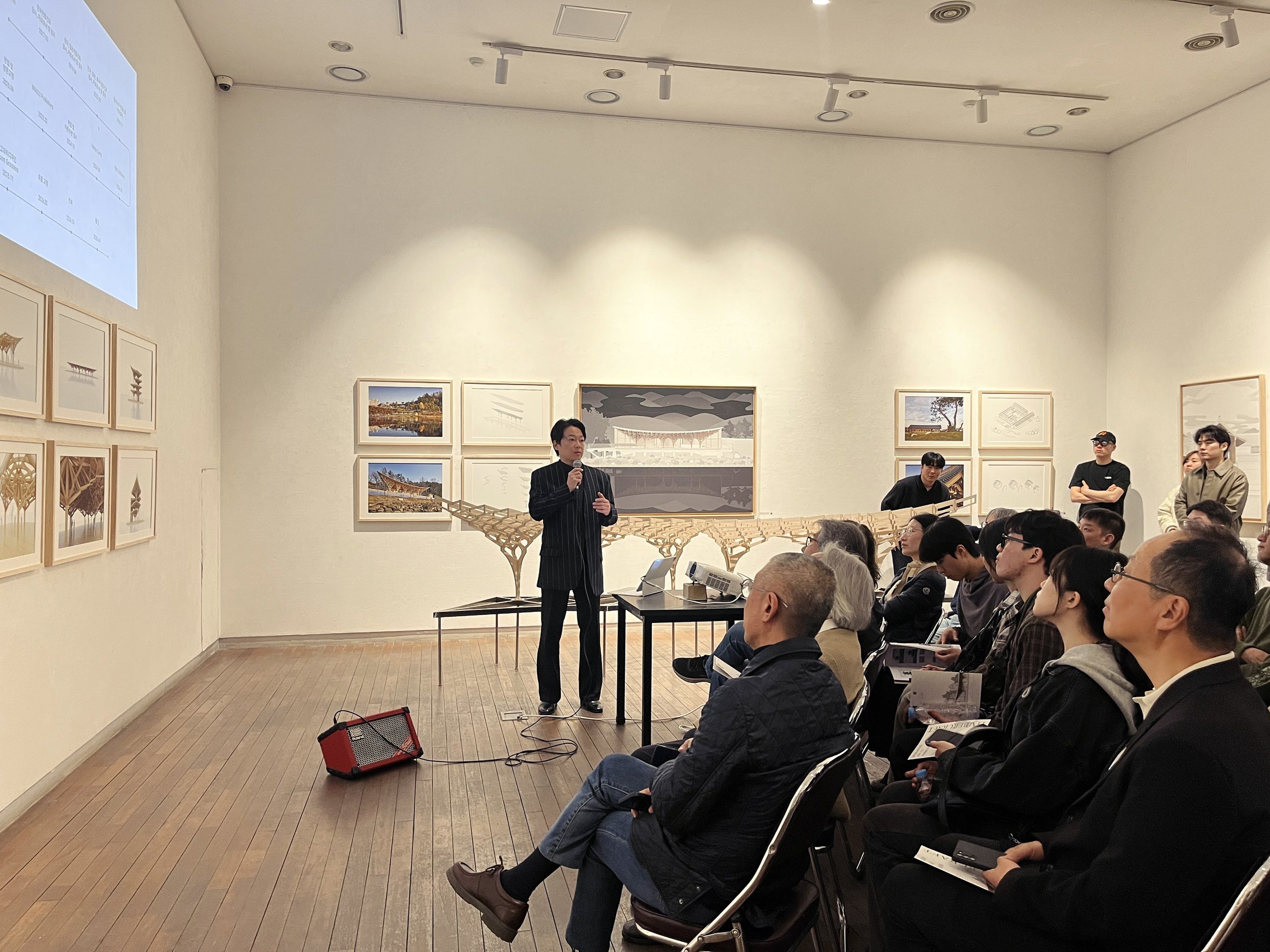 A man in a dark pinstripe suit is speaking into a microphone at a presentation or lecture. He stands in front of a table with a scale model of a building or structure. The audience is seated and attentively listening in a gallery or auditorium. The w