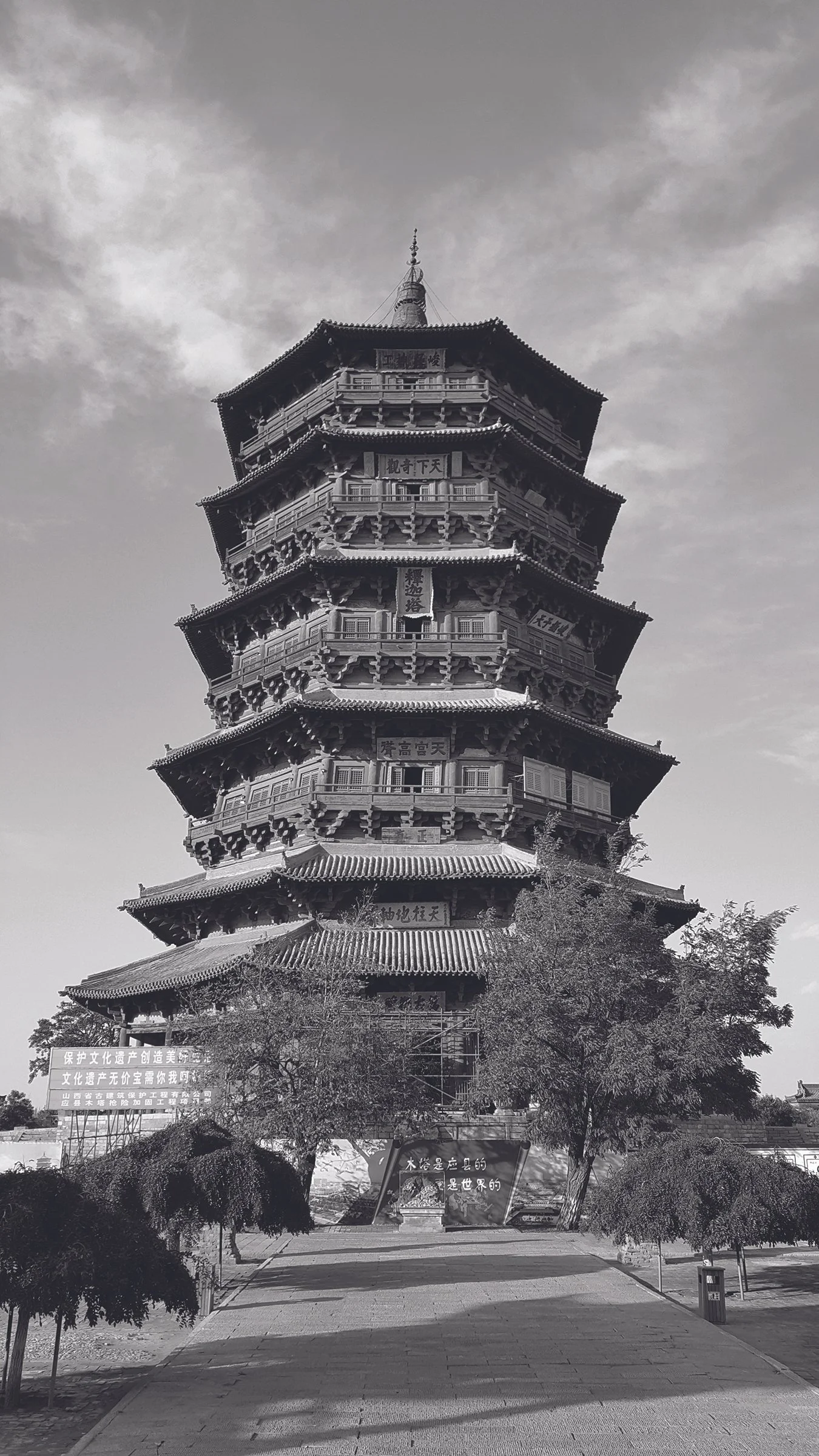 A traditional multi-story Chinese pagoda with ornate roofing and Chinese characters on plaques on each level, surrounded by trees under a cloudy sky.