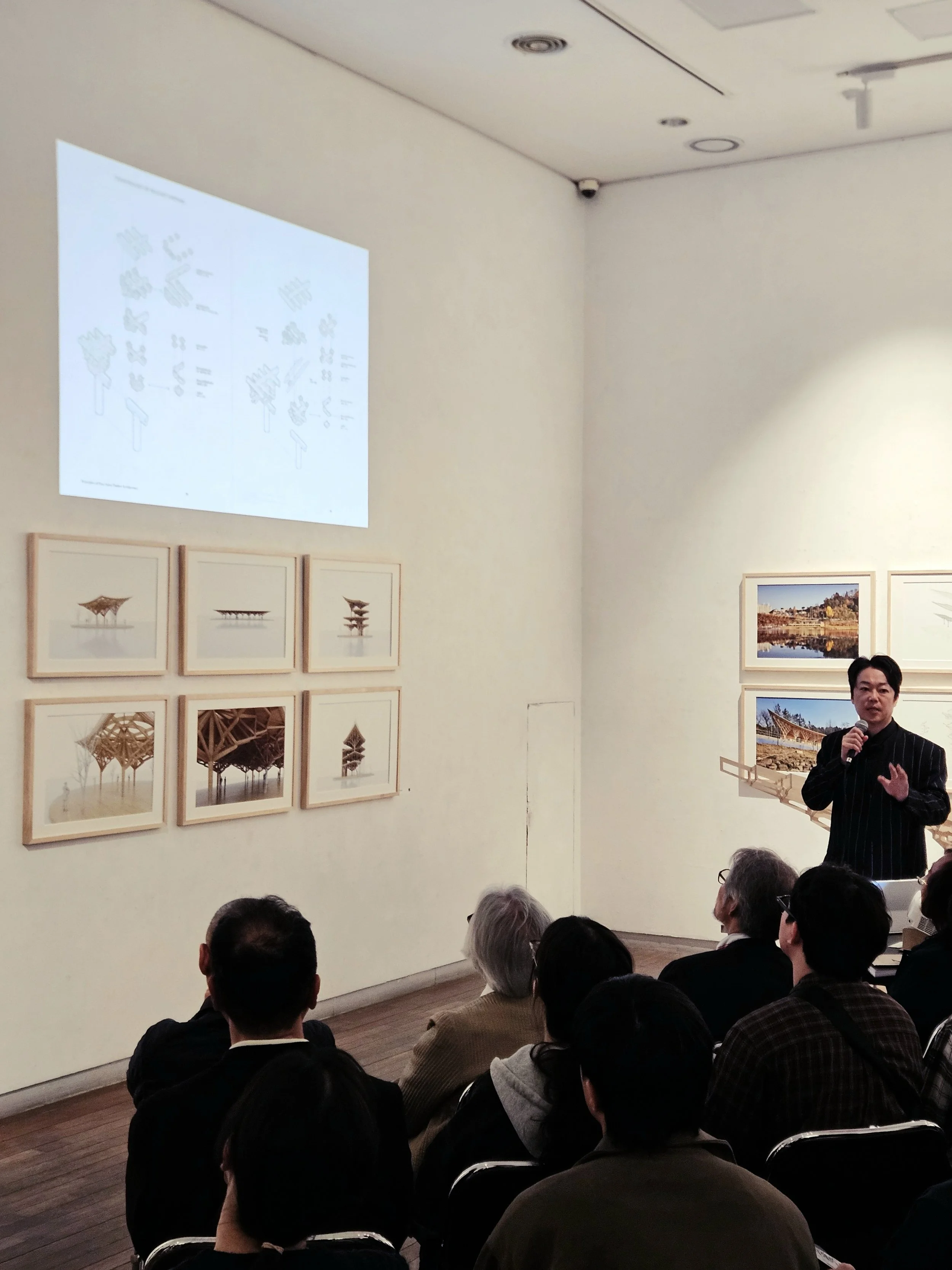A man in a black suit with white pinstripes holding a microphone, giving a presentation to an audience in an art gallery. The wall features framed photographs or artwork, and a diagram projected on the wall.
